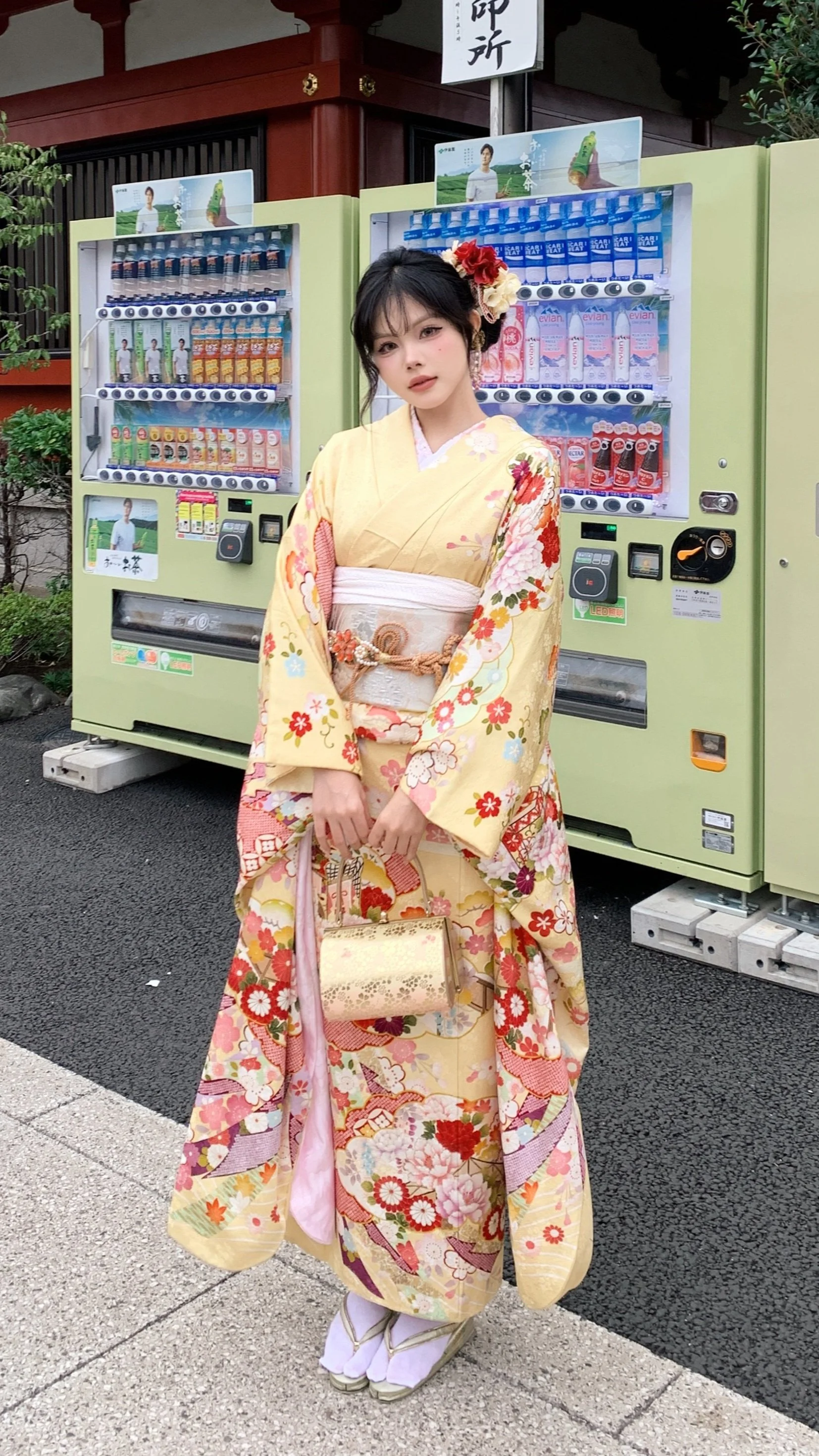 A young woman dressed in a traditional Japanese kimono with a floral pattern stands in front of a vending machine. She has a flower hair accessory and holds a small handbag, with her hands clasped in front of her.