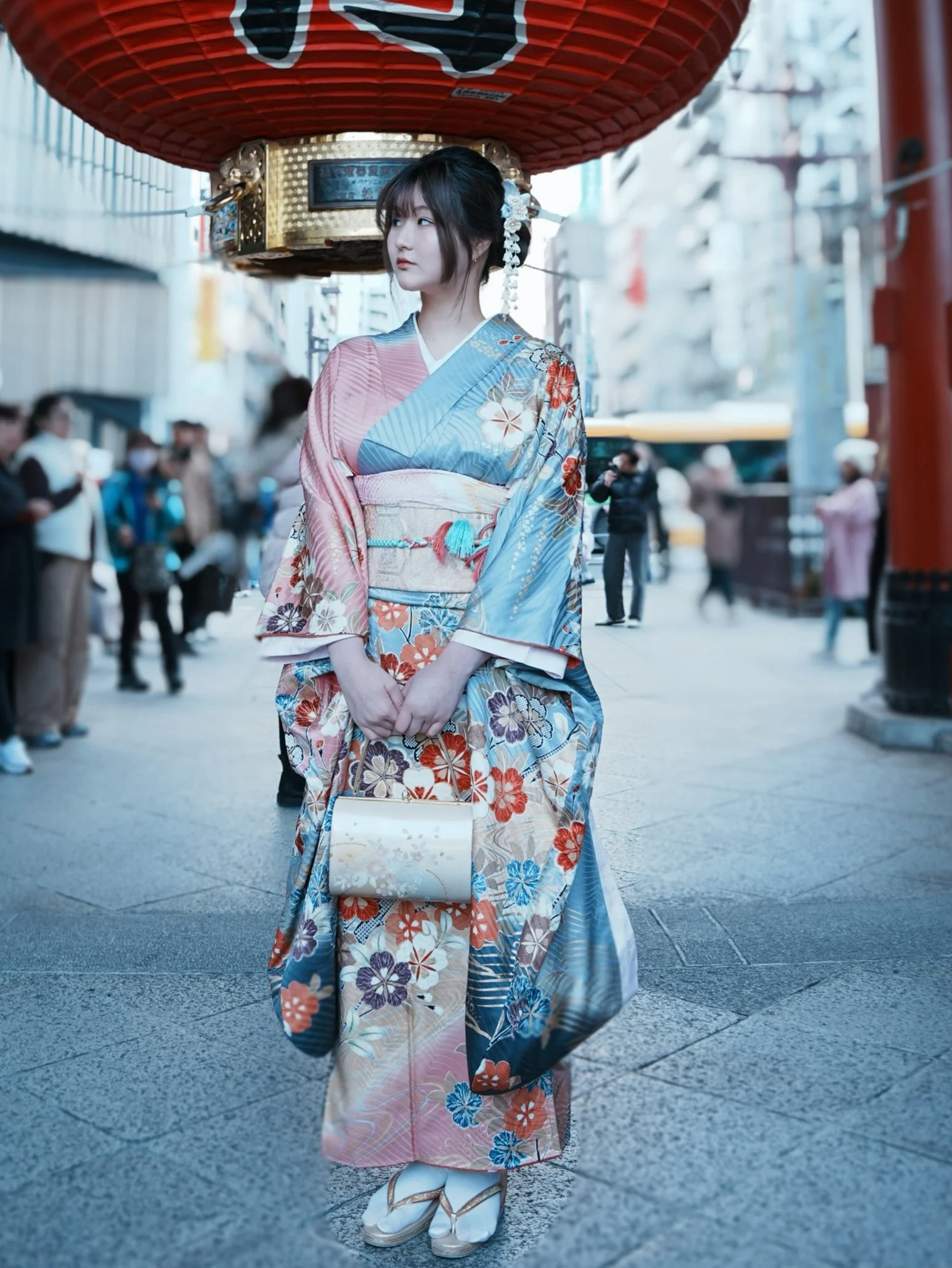A young woman wearing a colorful kimono standing under a large red paper lantern on a busy street.