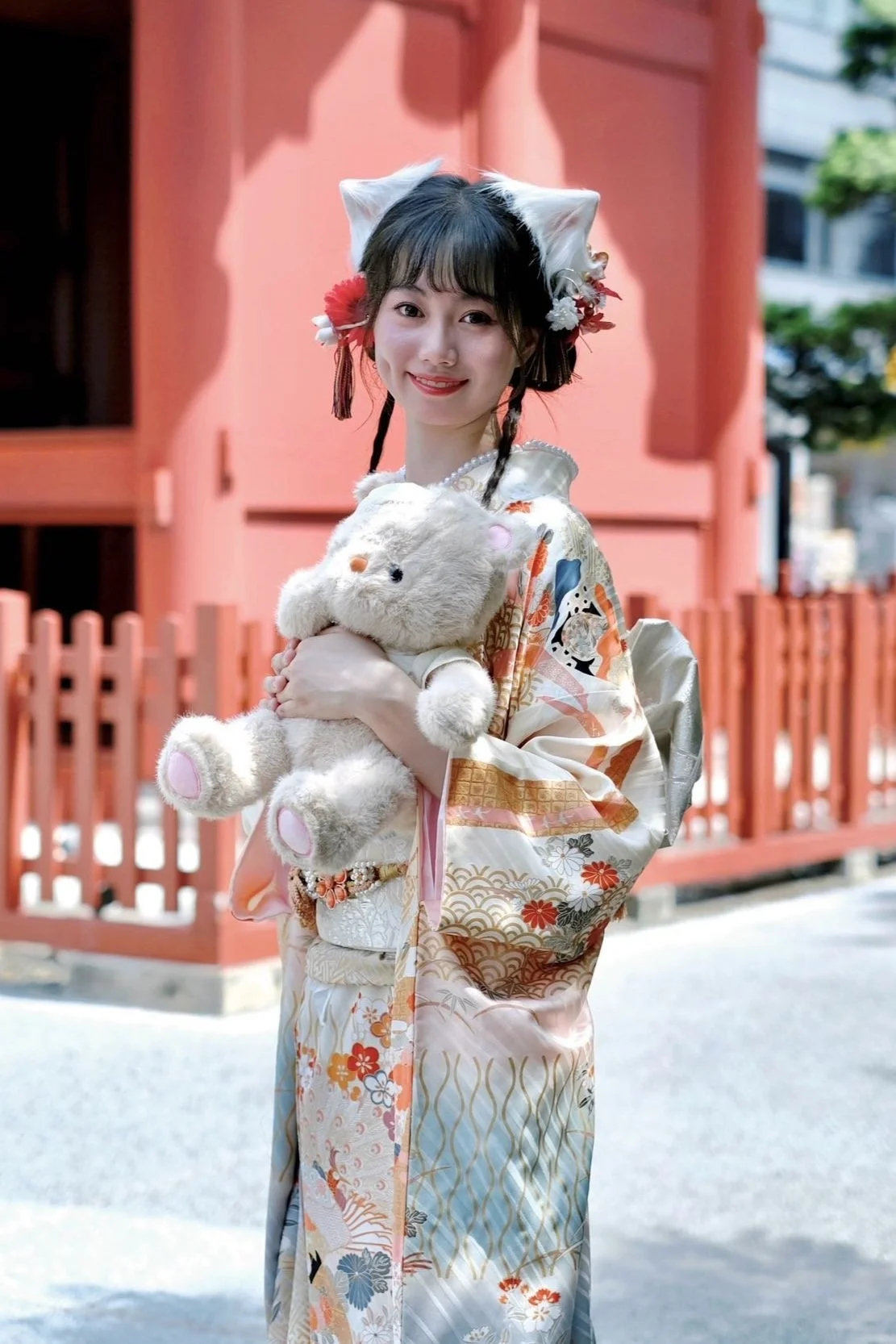 A young woman dressed in a traditional kimono holding a plush bunny toy, standing outdoors in front of a red fence with a building and trees in the background. She has cat ears on her head and is smiling at the camera.