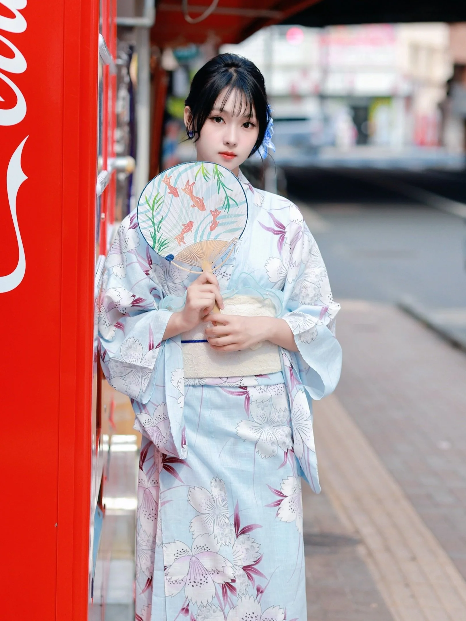 A young woman in a light blue floral yukata stands beside a red vending machine on a city street, holding a decorative fan with a koi fish and water lily design.
