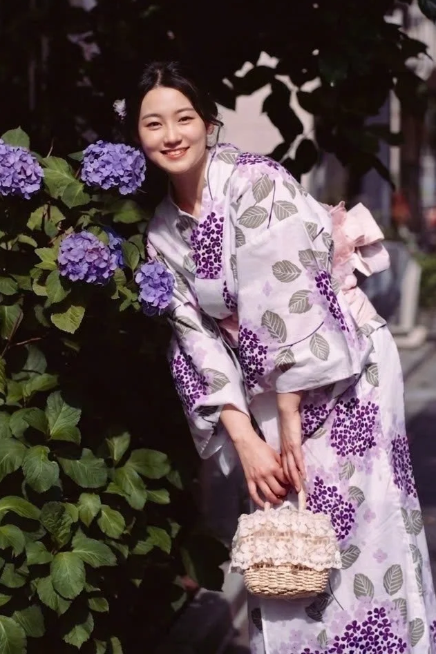 A mannequin dressed in a white yukata with purple and green floral patterns, a pink obi with a decorative flower clasp, and a lace bow, displayed in a shop window.