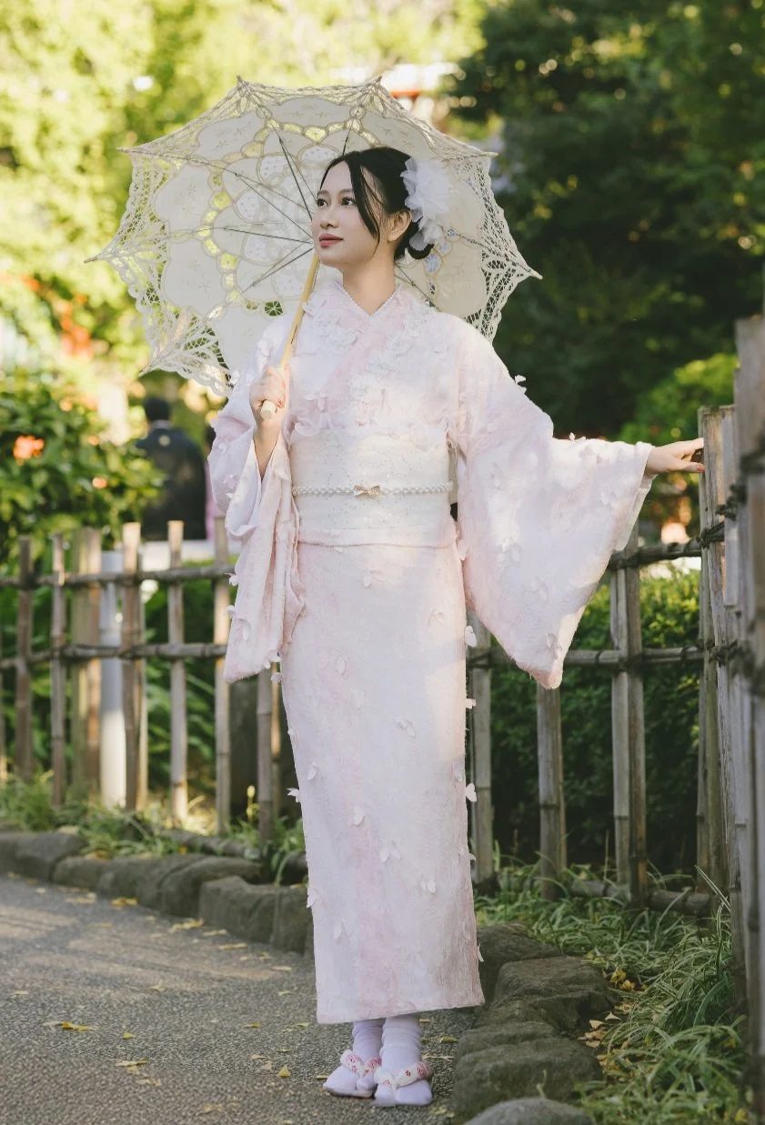 A woman dressed in a traditional light pink kimono with floral details, holding a lace parasol, standing outdoors near a wooden fence with greenery in the background.