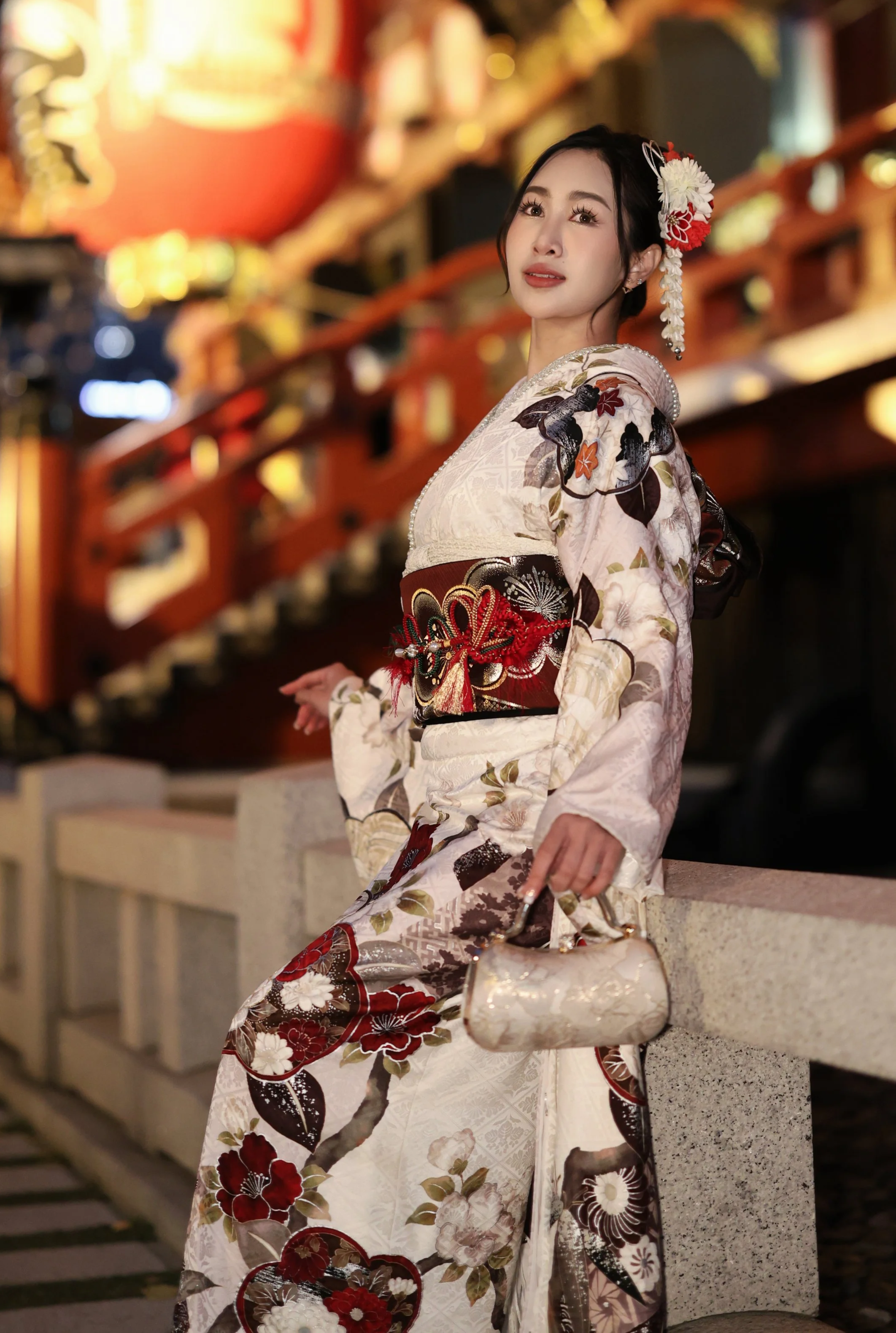 A young woman in a traditional Japanese kimono with floral patterns, holding a white handbag, standing near a red temple or shrine.
