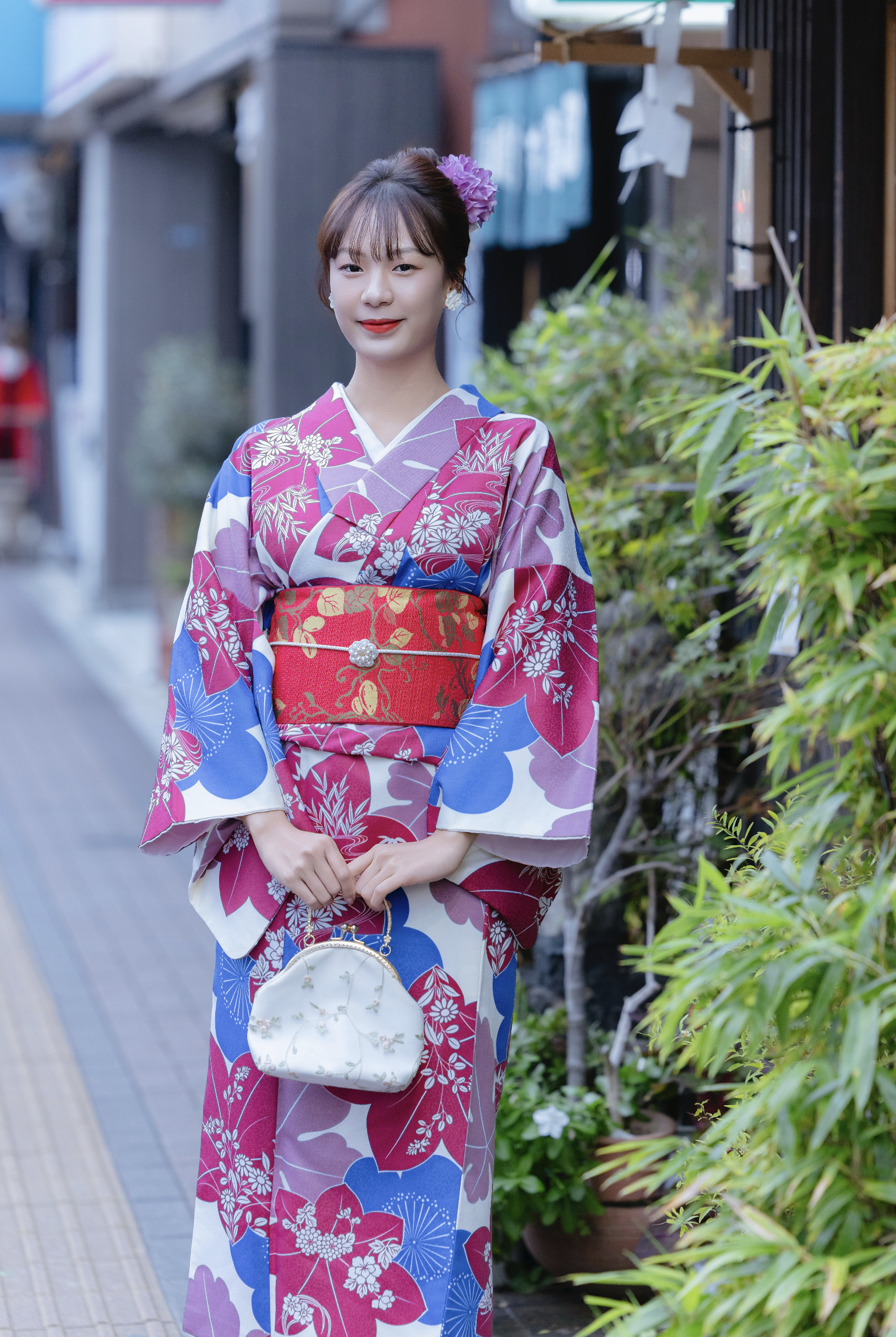 A woman wearing a colorful yukata with floral patterns, holding a small white handbag, standing on a sidewalk next to green plants.