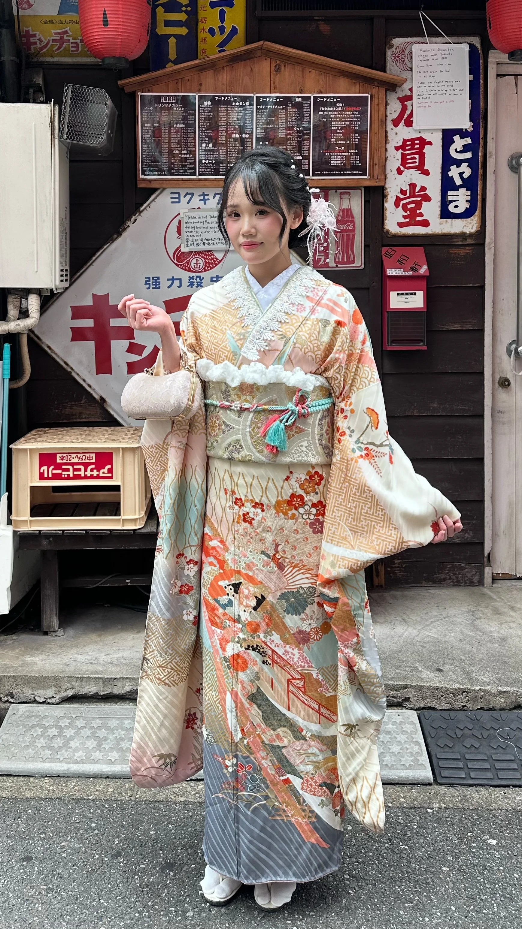 A young woman dressed in a traditional kimono holding a plush bunny toy, standing outdoors in front of a red fence with a building and trees in the background. She has cat ears on her head and is smiling at the camera.