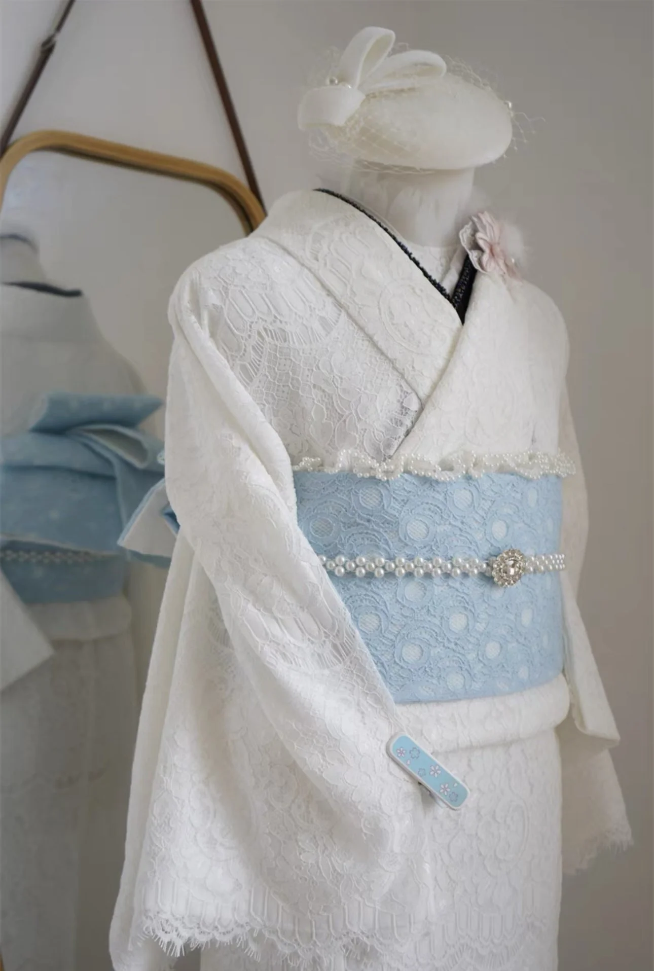 A Japanese doll dressed in a traditional white kimono with light blue obi, adorned with pearl embellishments, standing near a mirror and a box with fabric accessories.
