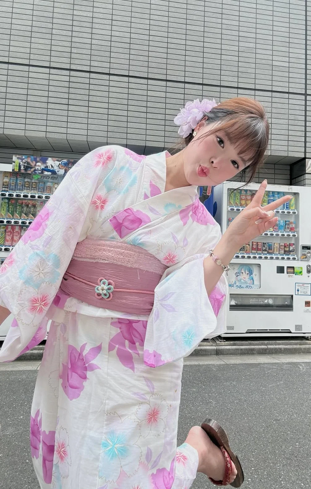 A mannequin dressed in a pastel-colored floral kimono with a pink obi and a white decorative cord. The mannequin wears a white fox mask decorated with pink floral patterns, red tassels, and red ears, with the mask covering the face.