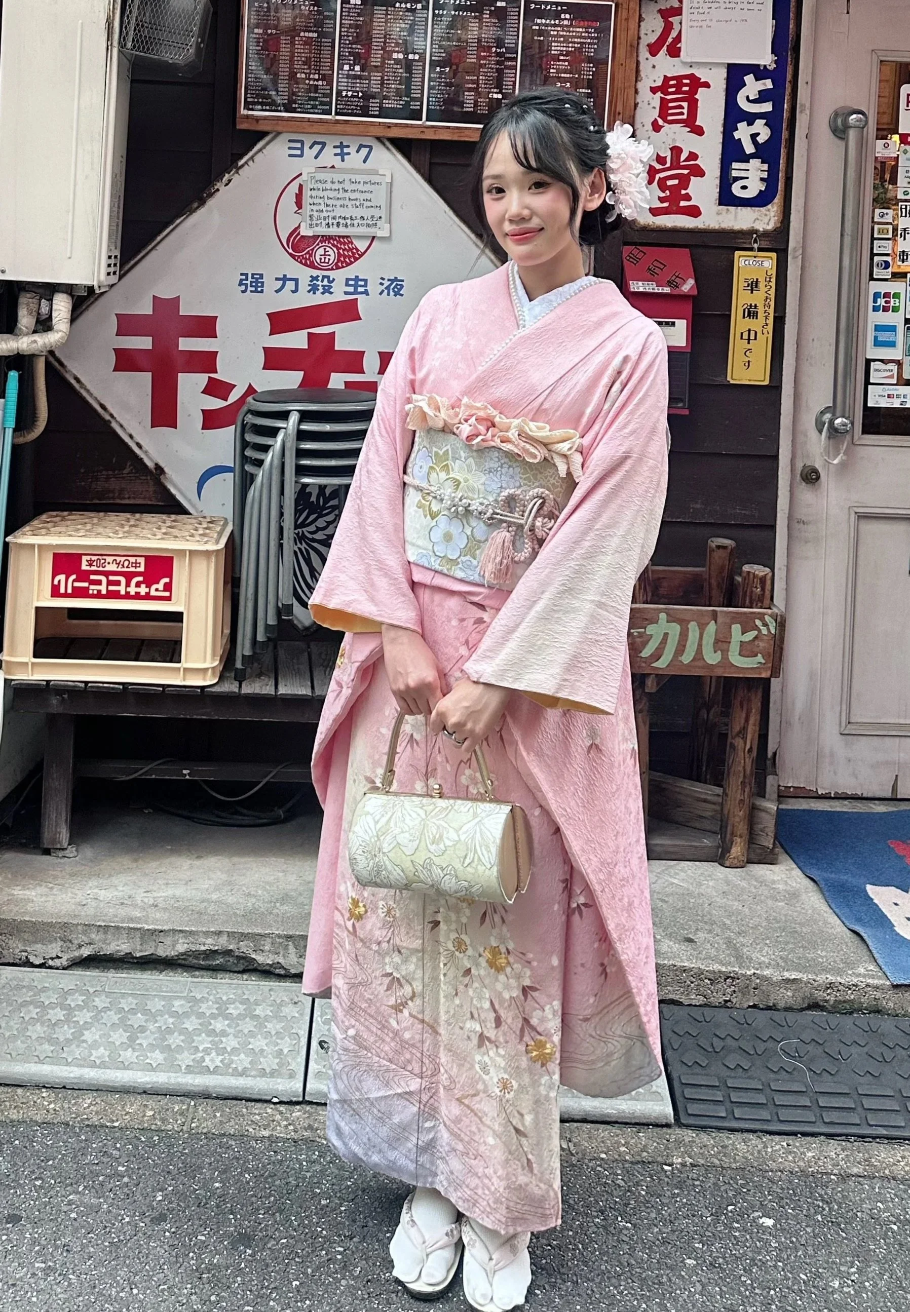 A woman dressed in a pink kimono with floral patterns, standing in front of a traditional Asian temple or shrine.