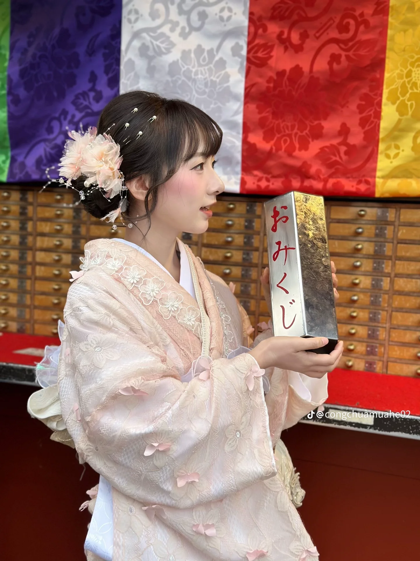 A woman dressed in a traditional light pink kimono with floral details, holding a lace parasol, standing outdoors near a wooden fence with greenery in the background.