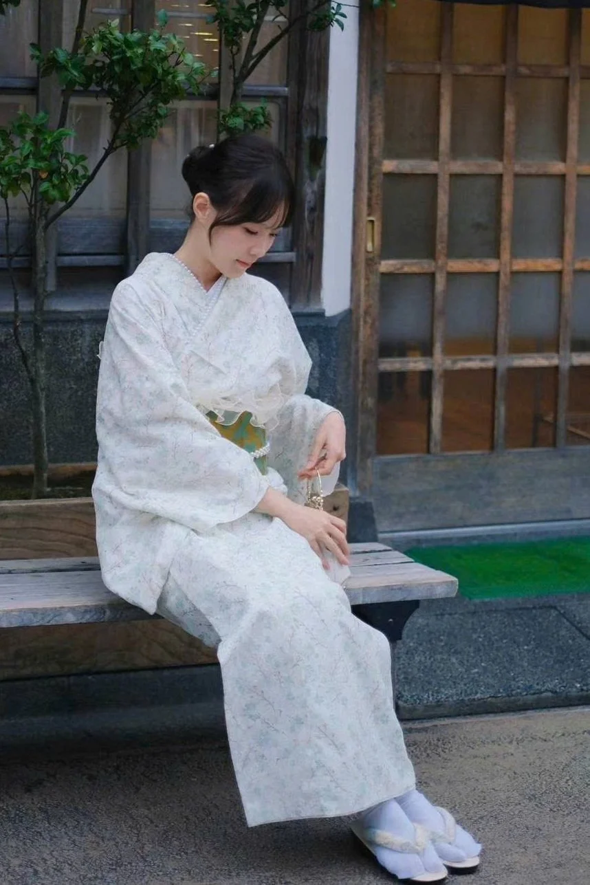 A young woman dressed in a traditional white kimono sitting on a wooden bench outdoors, holding a small purse, with a background of wooden elements, plants, and stone.