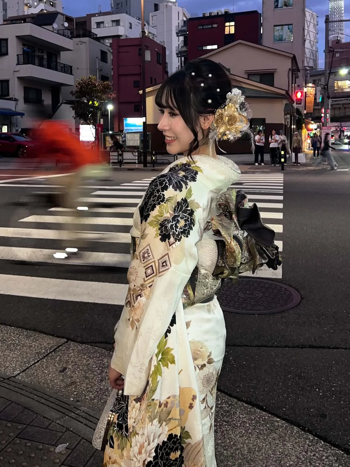 Young woman in traditional Japanese kimono holding a paper parasol. She has dark hair with a hair ornament, standing outdoors near red and black structures.