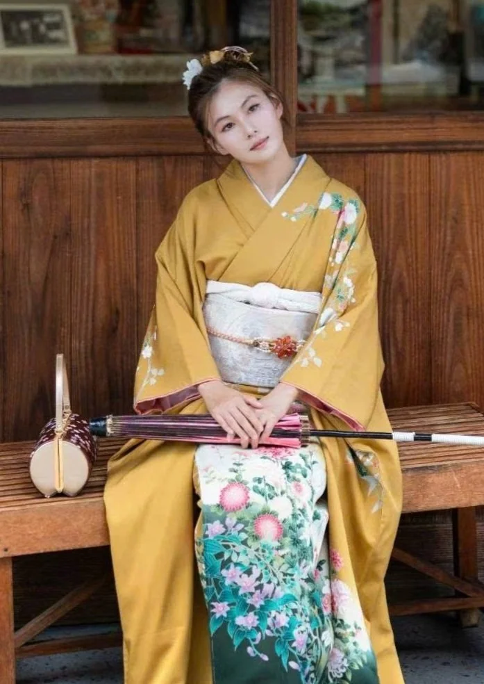 A woman wearing a traditional Japanese kimono with floral designs, sitting on a wooden bench, holding an umbrella, and posed with her head tilted.