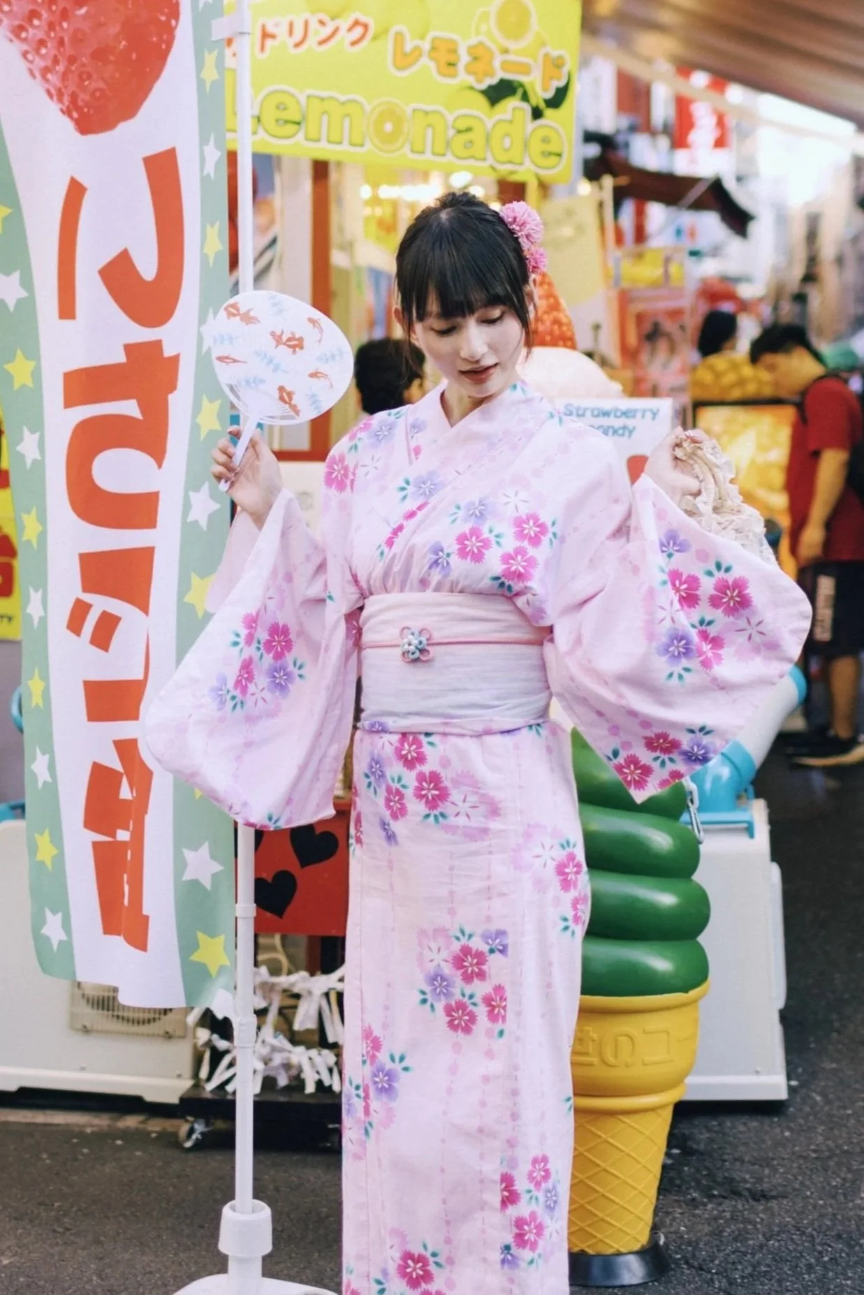 A young woman wearing a pink floral yukata standing at a street festival, holding a small fan, with colorful signs and food stalls in the background.