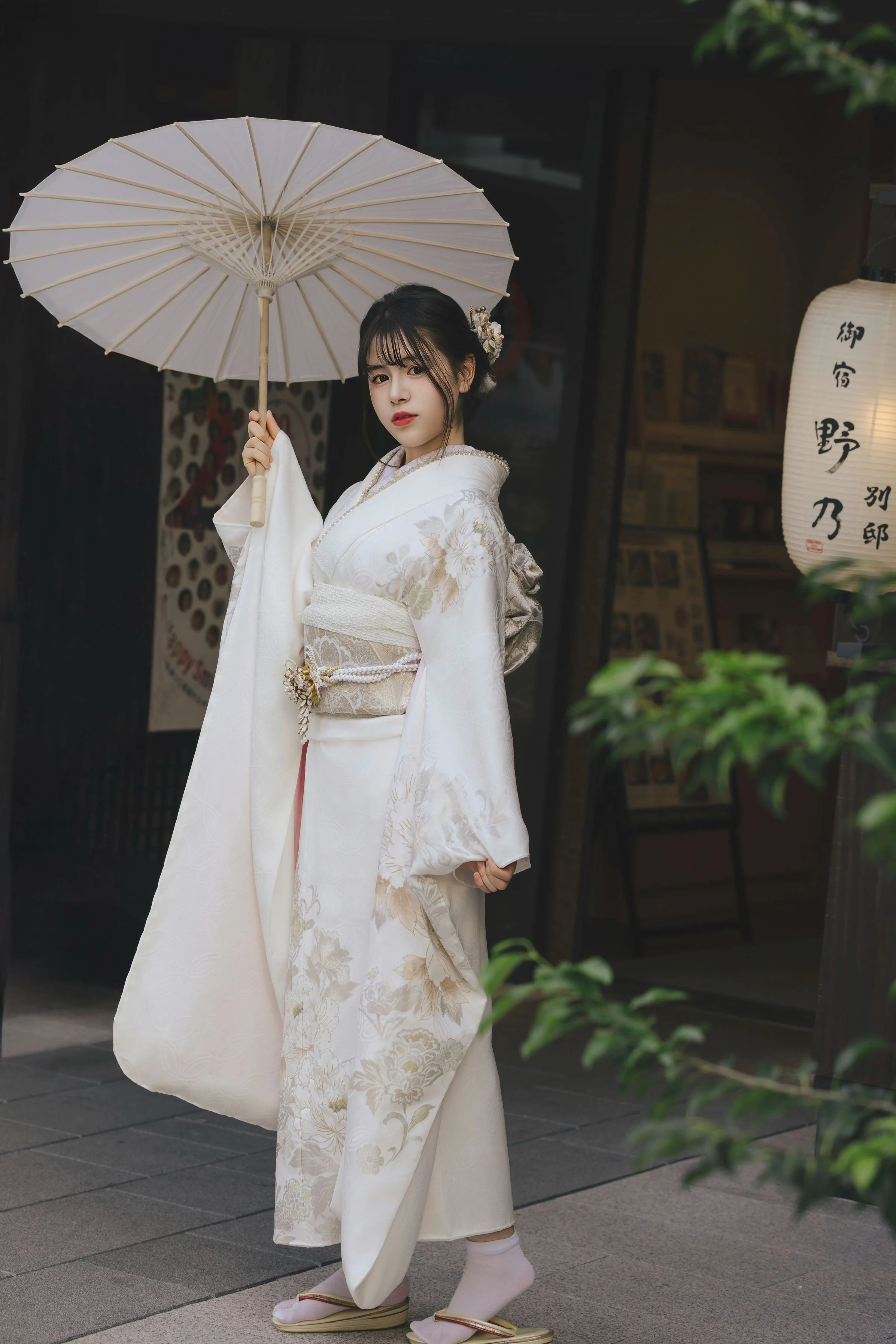 A young woman wearing a traditional white kimono with floral patterns, holding a paper umbrella, standing outside a building in Japan.