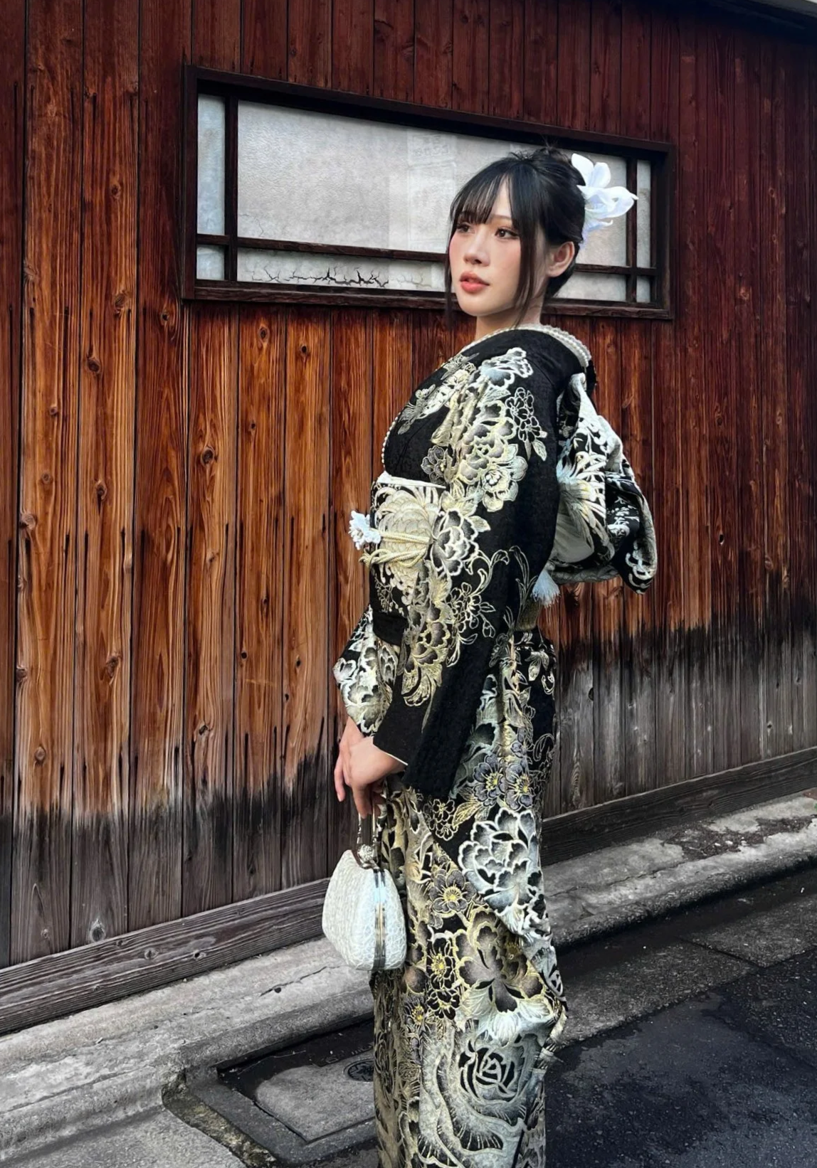A young woman in a traditional black and white kimono with floral patterns, standing outdoors against red-painted wooden structures, holding a small handbag.