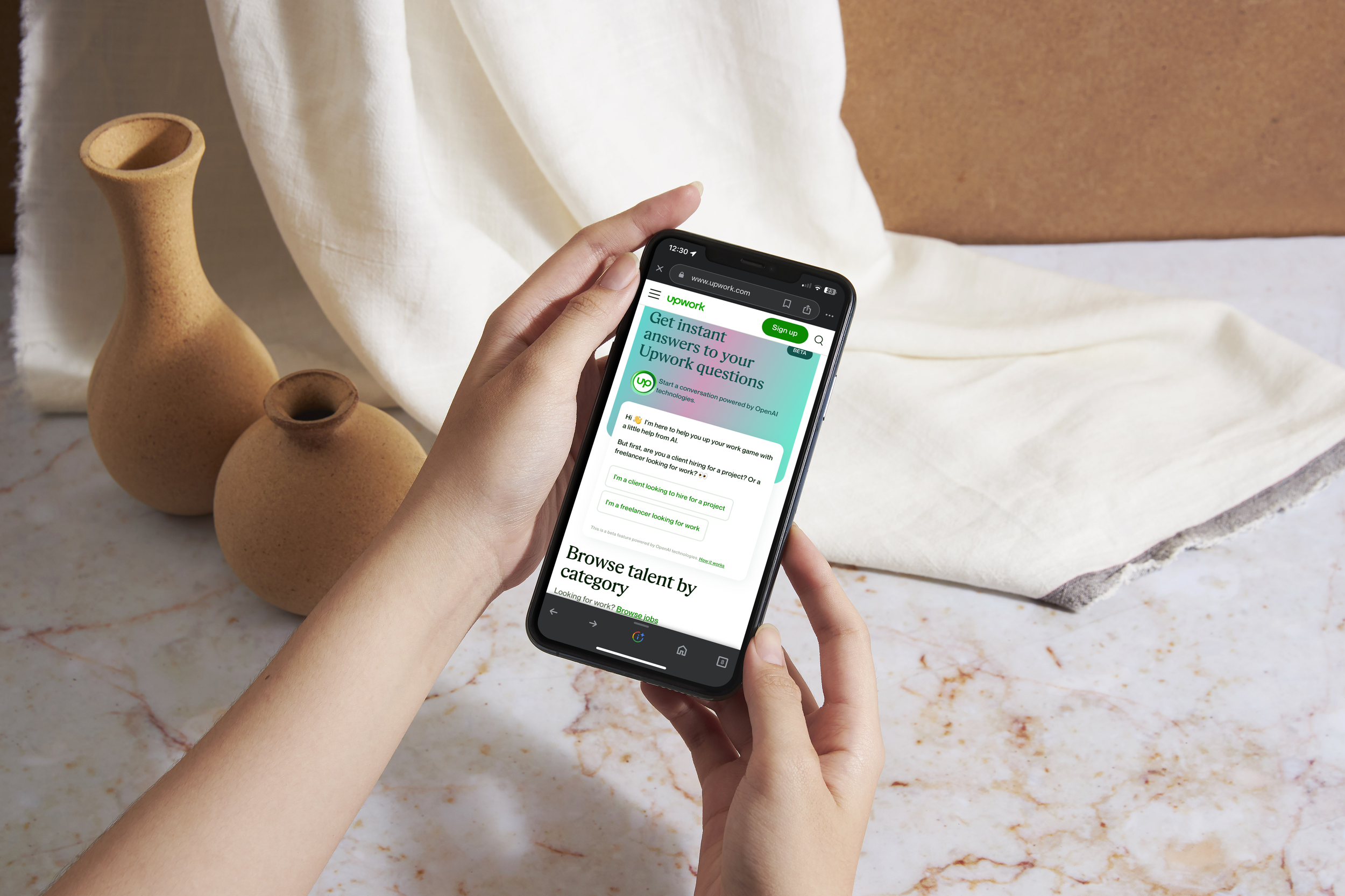 Person holding a smartphone displaying Upwork website on a marble countertop with beige vases and a white cloth backdrop.