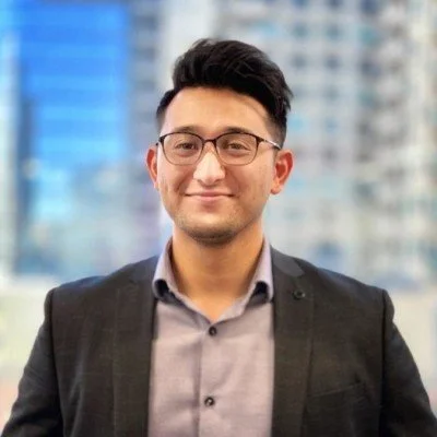 A man in a gray shirt and black blazer standing outdoors with a city skyline in the background.