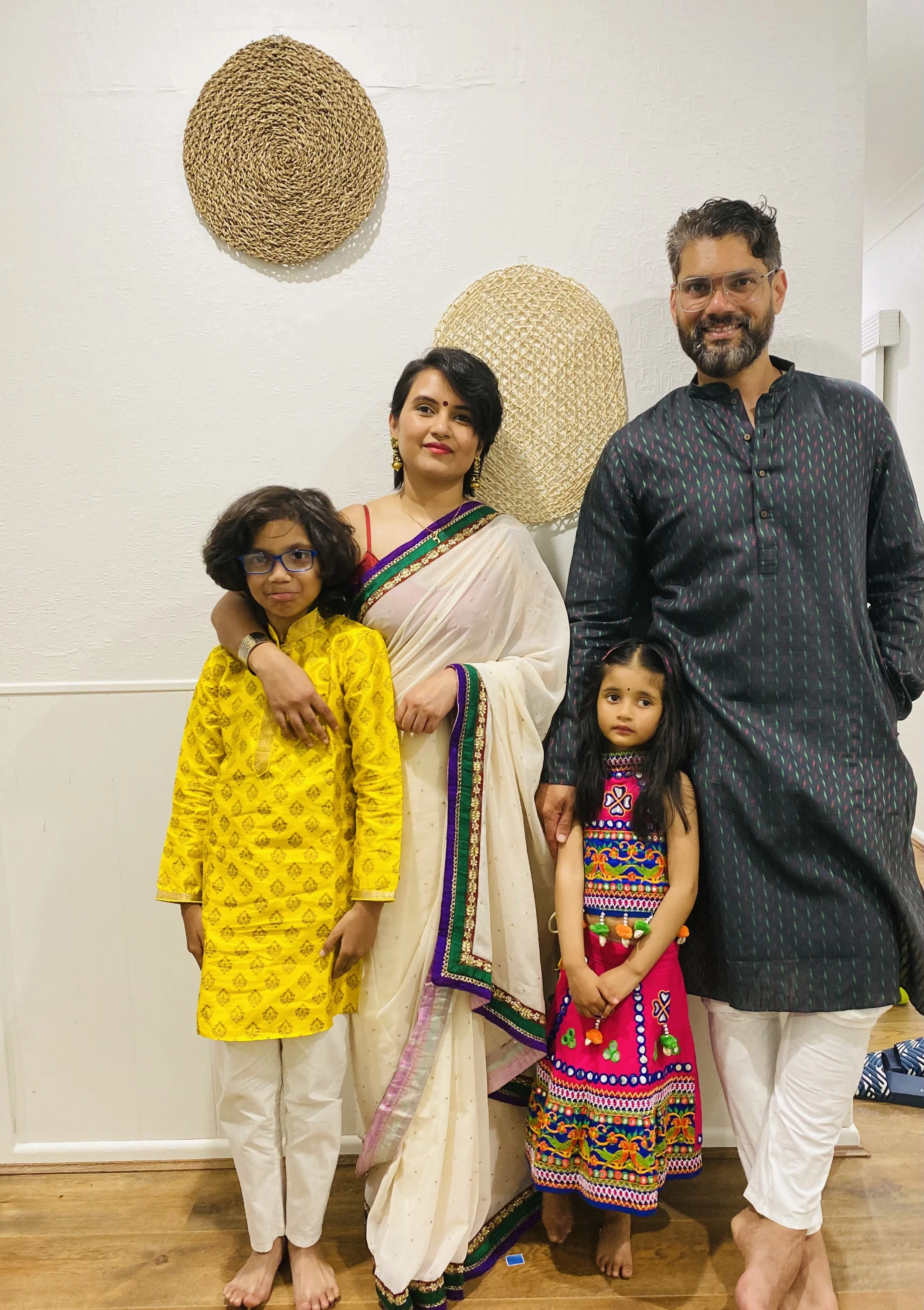 A multigenerational Indian family standing together indoors. The woman in the center wears a cream saree with colorful border; the man on the right wears a dark kurta with white pants. The three children are in traditional clothing: a girl in yellow, a girl in a multicolored dress, and a boy in a yellow kurta with glasses. All are smiling, with the children standing close to the adults.