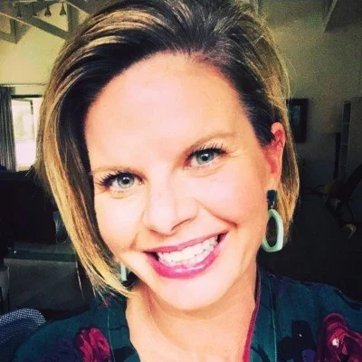 A smiling woman with short brown hair, wearing earrings and a dark floral top, indoors with a window in the background.