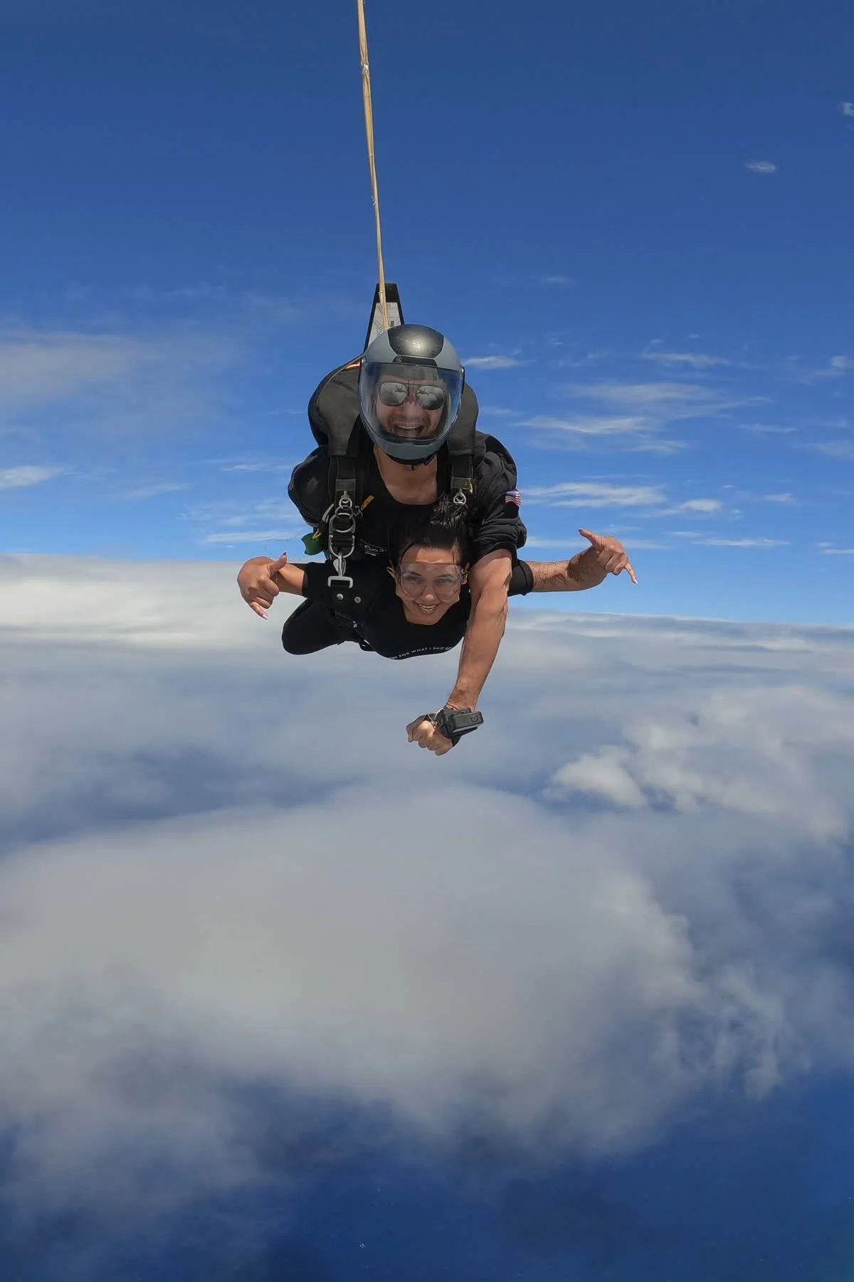 Two people in tandem freefall during a skydiving jump, smiling and making hand gestures, with blue sky and clouds below.