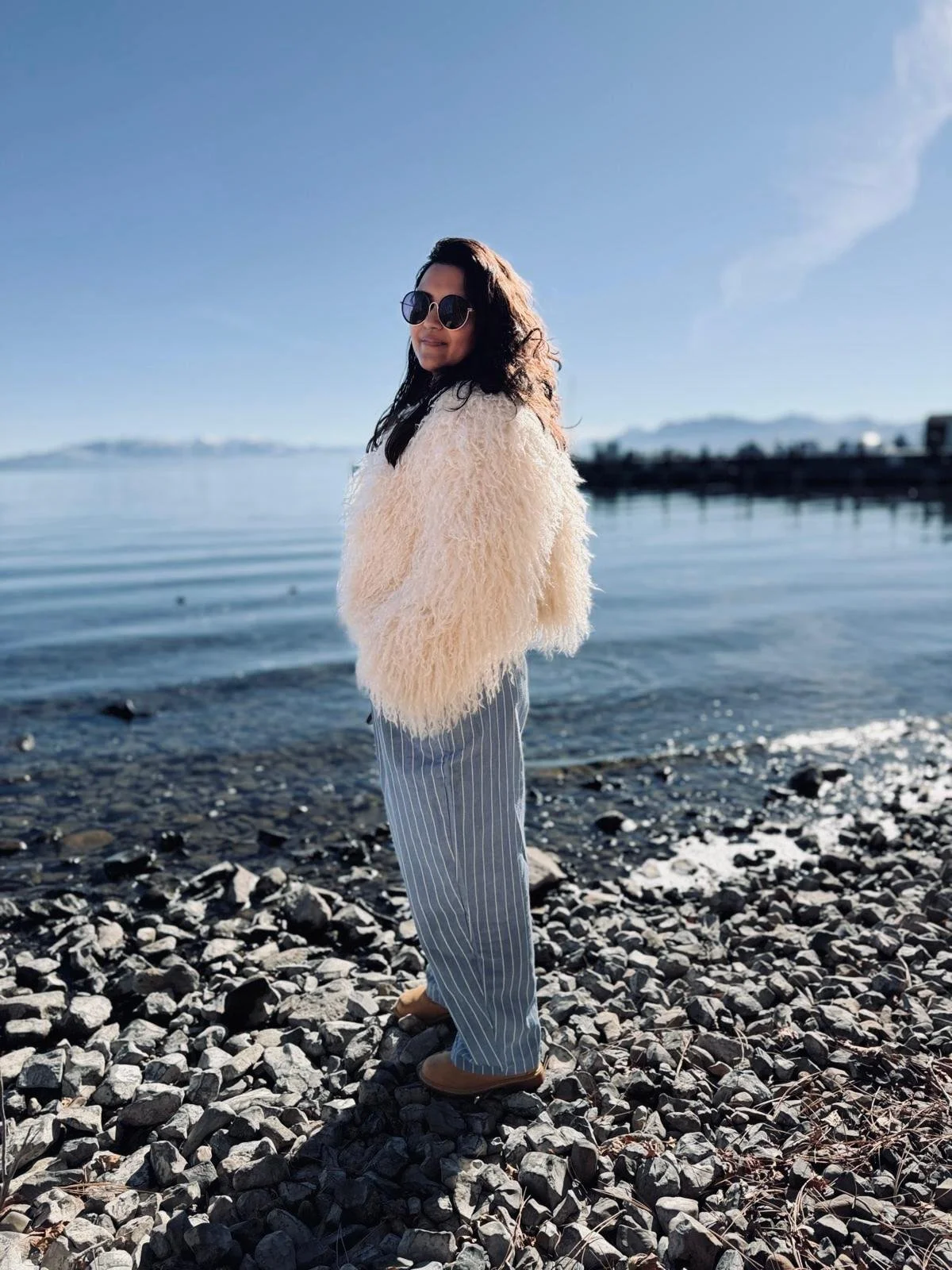 Woman standing on rocky shoreline by the water, wearing a fluffy beige jacket, striped pants, brown shoes, sunglasses, and looking at the camera with a smile, with clear blue sky and distant mountains in the background.