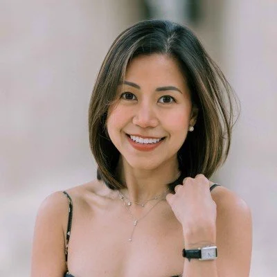 A young woman with shoulder-length dark hair smiling, wearing a black spaghetti strap top, a necklace, earrings, and a watch, outdoors with a blurred background.