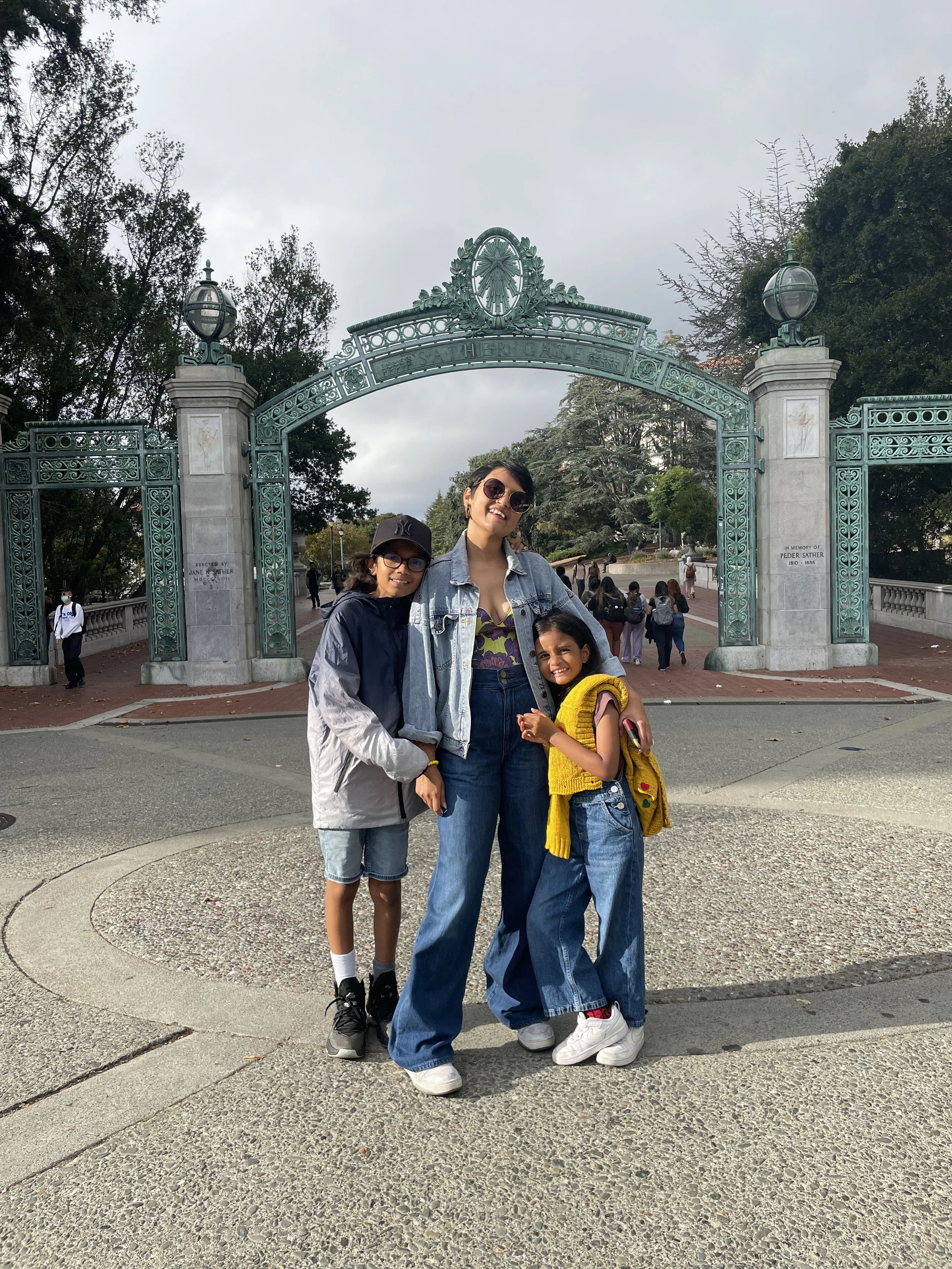 Three people posing in front of the Sather Gate entrance at UC Berkeley on a cloudy day, surrounded by trees and other visitors.