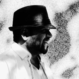 A black and white photo of a man smiling, wearing a fedora hat and a white shirt, with a textured background.