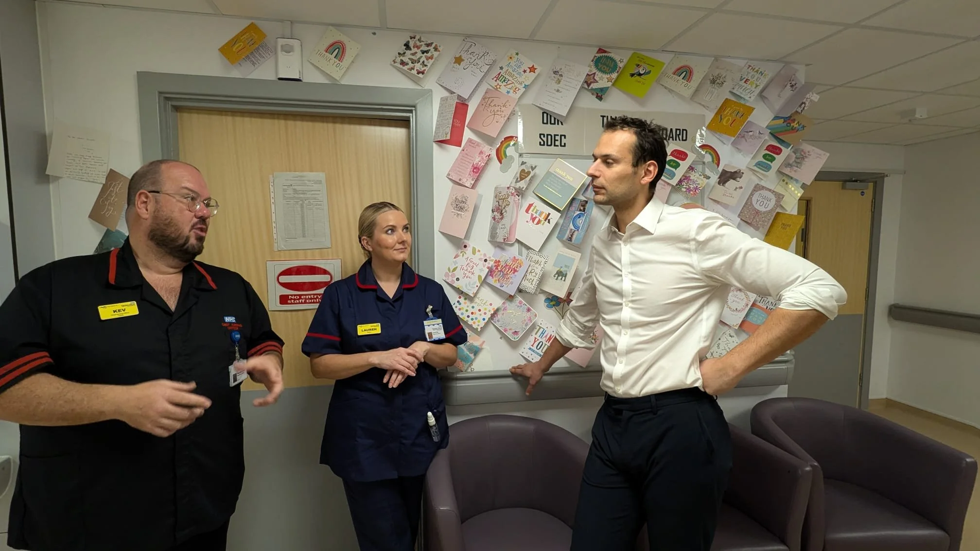 Josh Simons MP in a white shirt talking to two nurses in navy uniforms inside a hospital, with birthday and thank you cards displayed on the wall behind them.