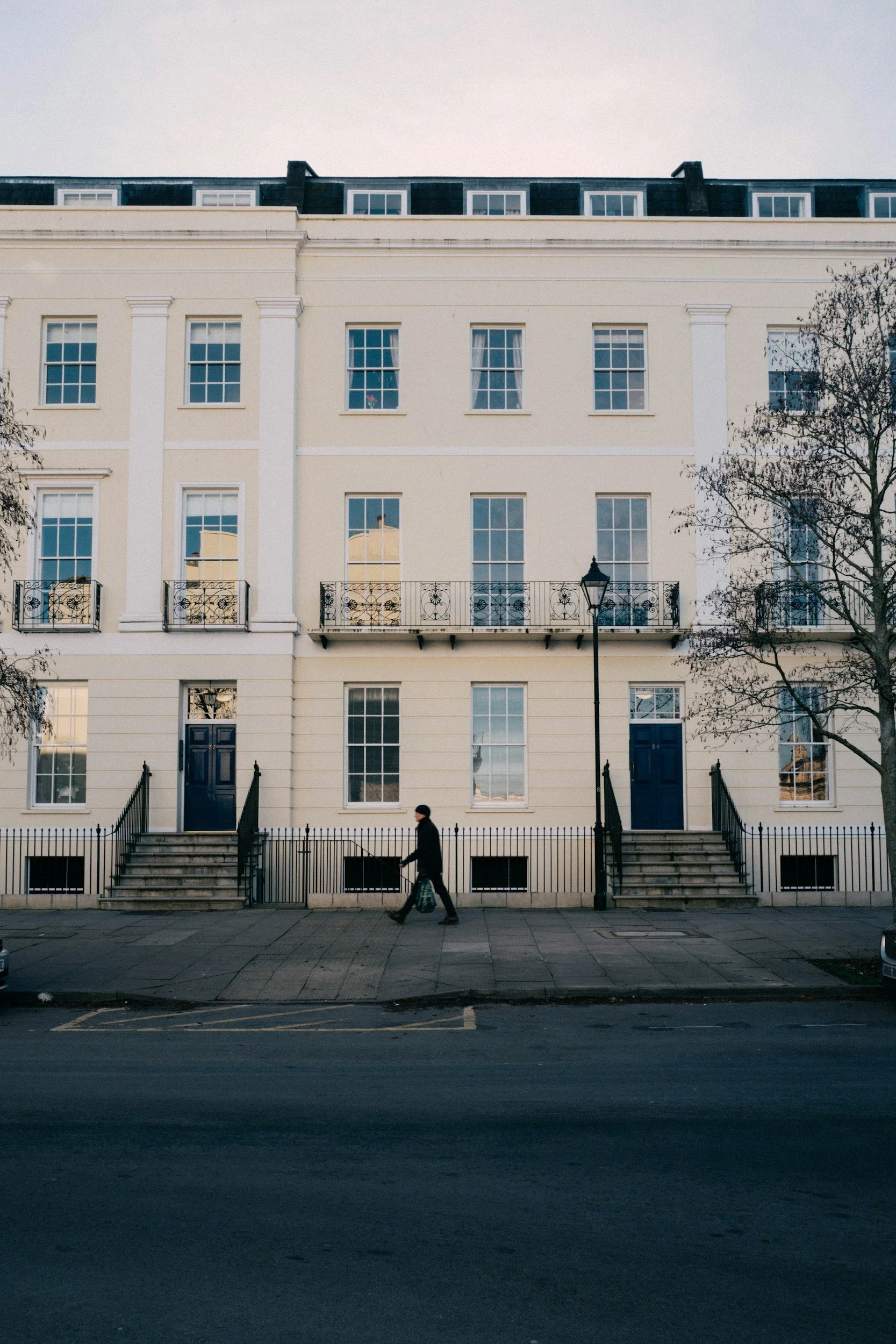 A person in dark clothing walking past a white, multi-story building with stairs leading to dark blue doors and several large windows. There are railings, a streetlamp, trees, and parked cars visible.