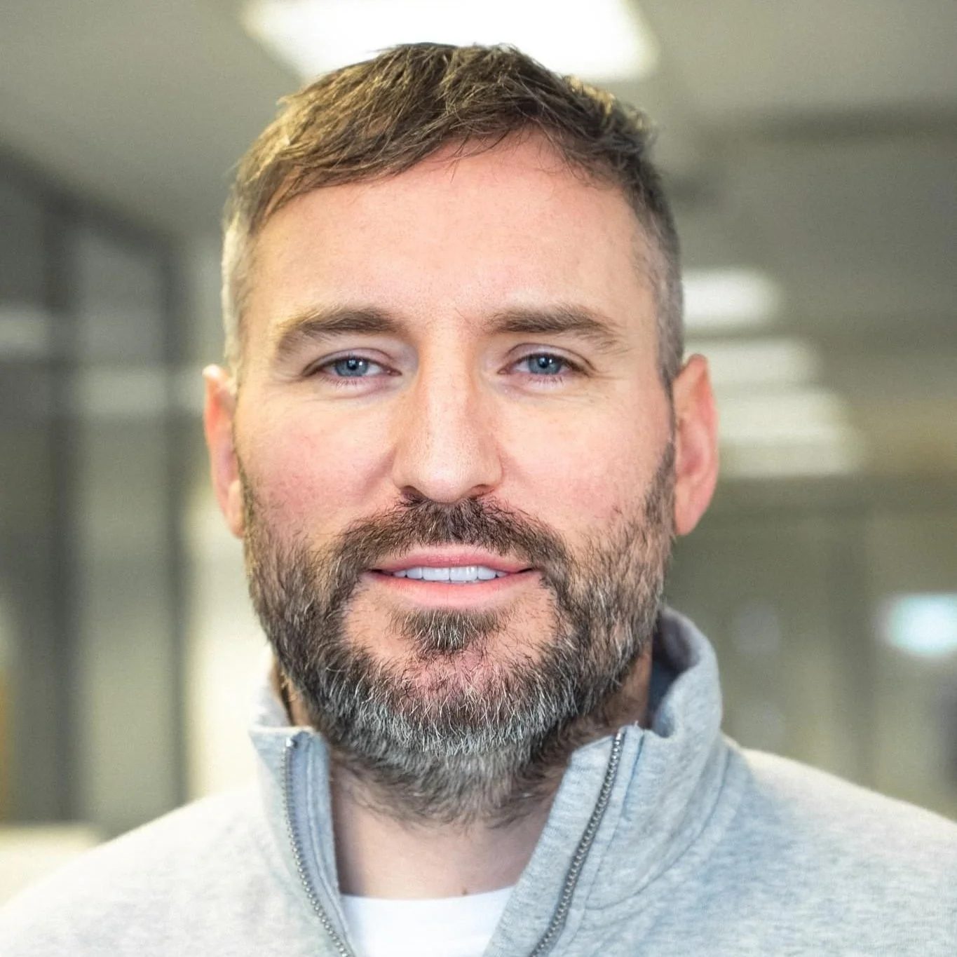 A man with short brown hair, blue eyes, and a beard and mustache, wearing a gray quarter-zip fleece, smiling in an indoor environment.