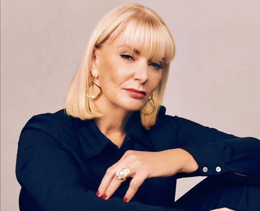 A woman with blonde hair and bangs, wearing a black shirt, gold hoop earrings, and a large ring, posing with her arm resting on her knee against a plain background.