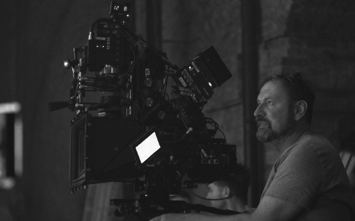 A man with a beard and short hair operating a professional film camera on a film set with a dark, industrial background.