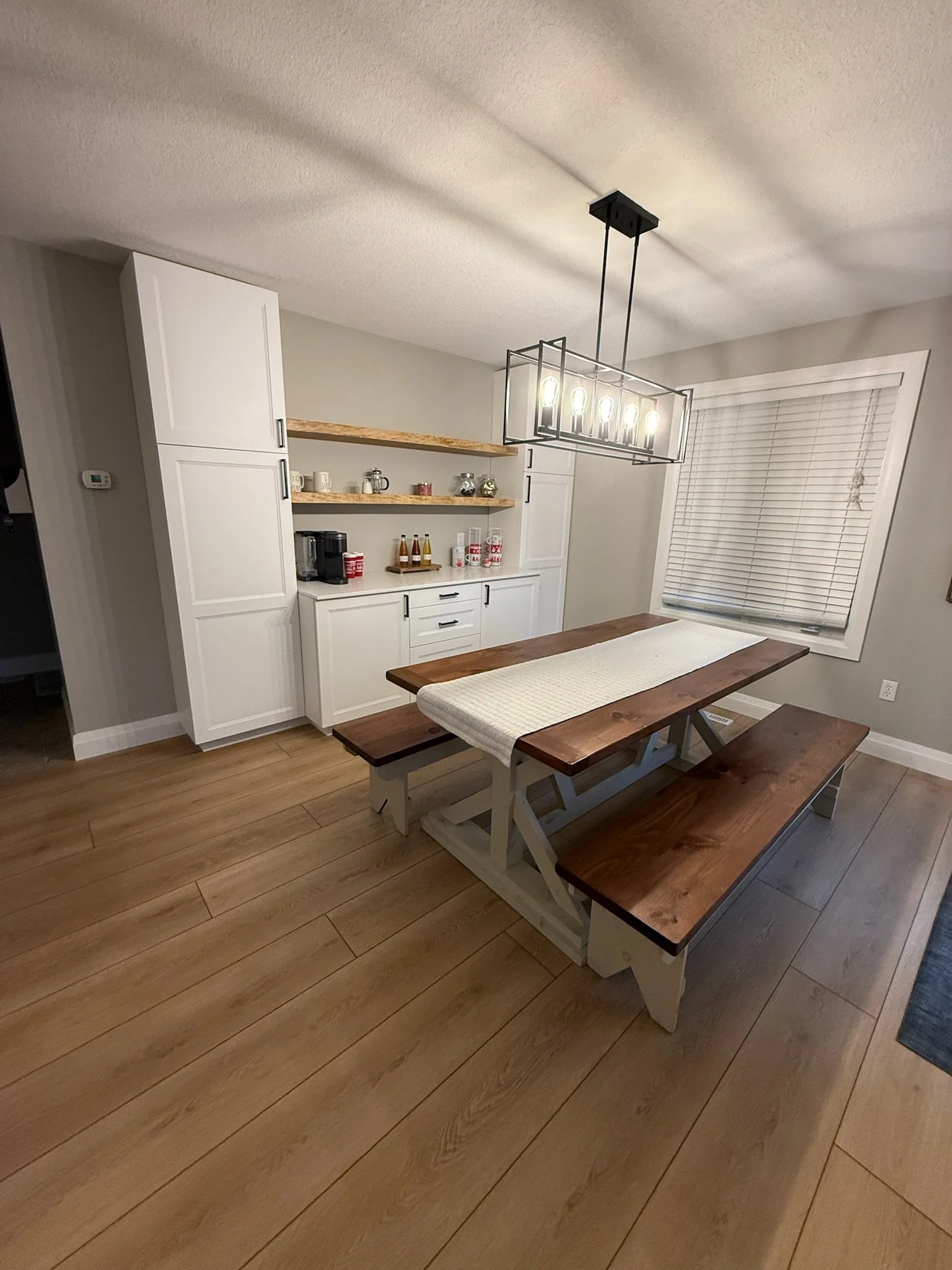 A dining room with a wooden table and matching benches, a white built in bar and pantry with open shelves filled with mugs and bottles, a black chandelier with exposed bulbs, a window with closed blinds, and light wood flooring.