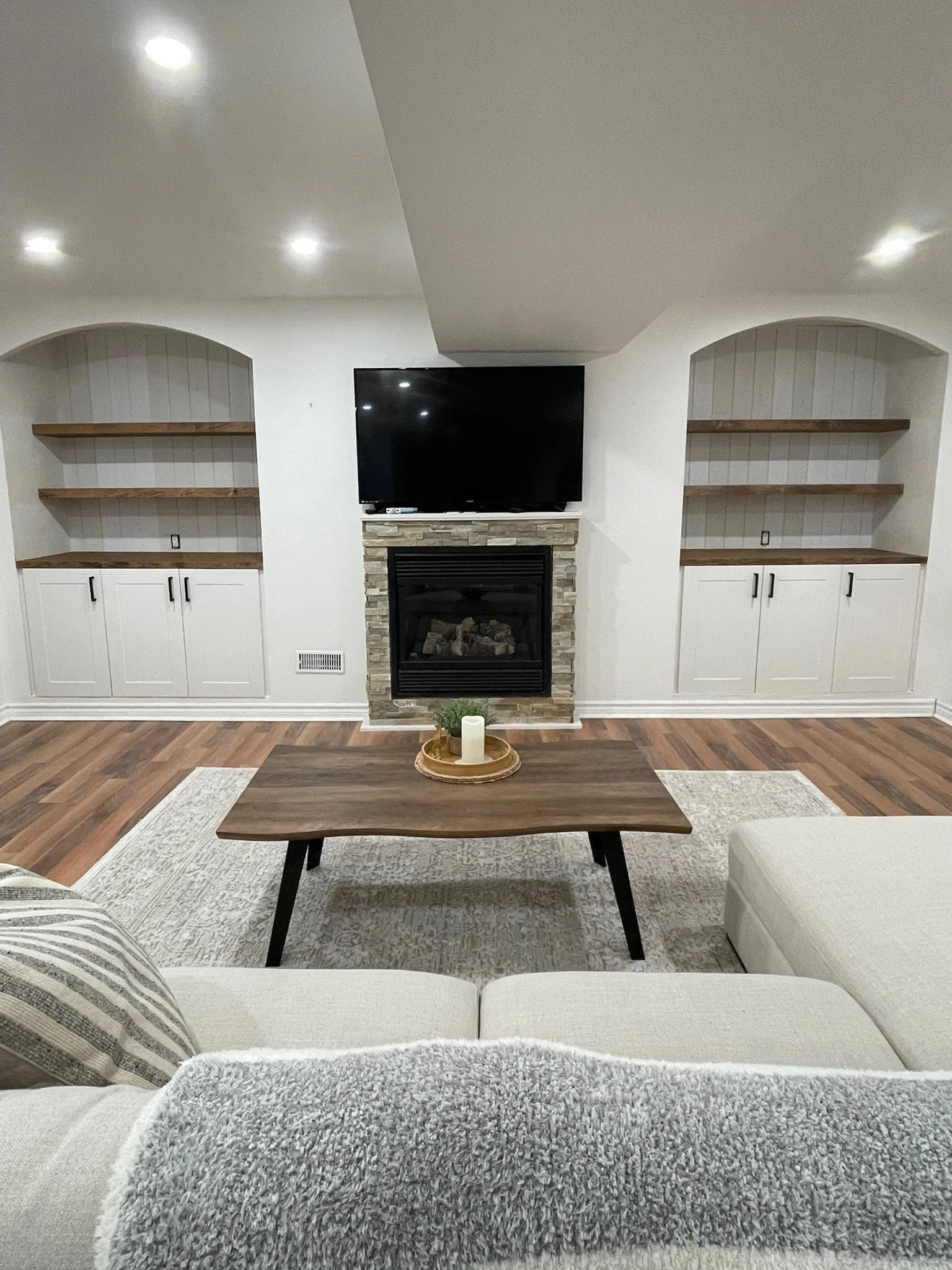 Living room with a beige sofa, a wooden coffee table, a decorative tray with candles and plants, a fireplace with a black screen, a wall-mounted TV, and built-in shelves and arched trim detail on either side of the fireplace.
