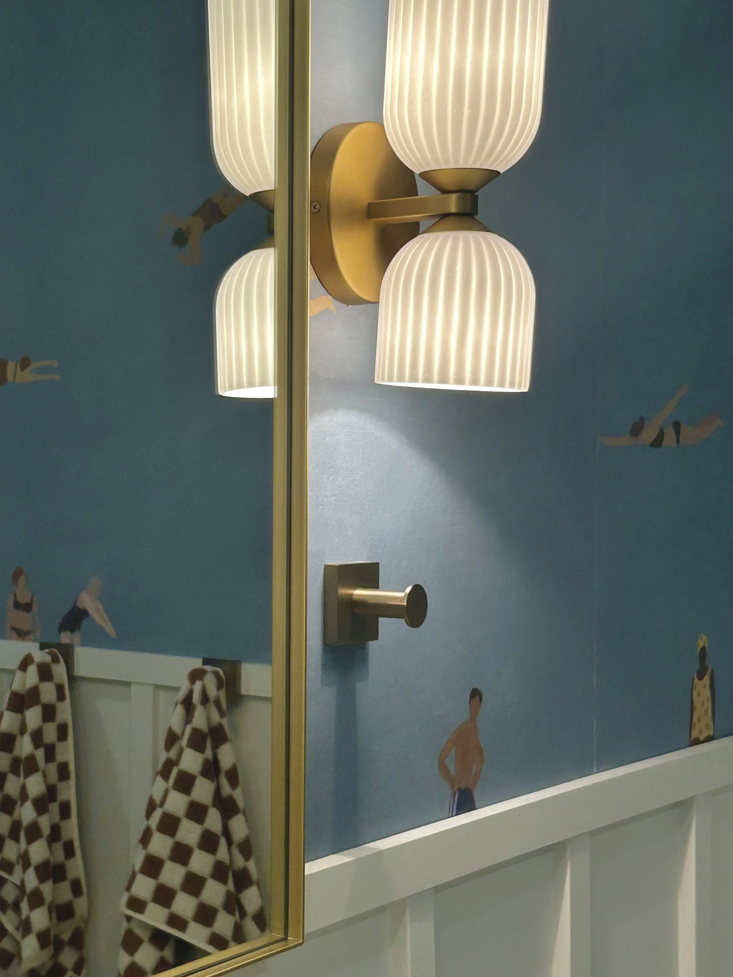 A wall-mounted light fixture with three ribbed glass shades illuminated, against a blue wall decorated with ballet dancer illustrations.