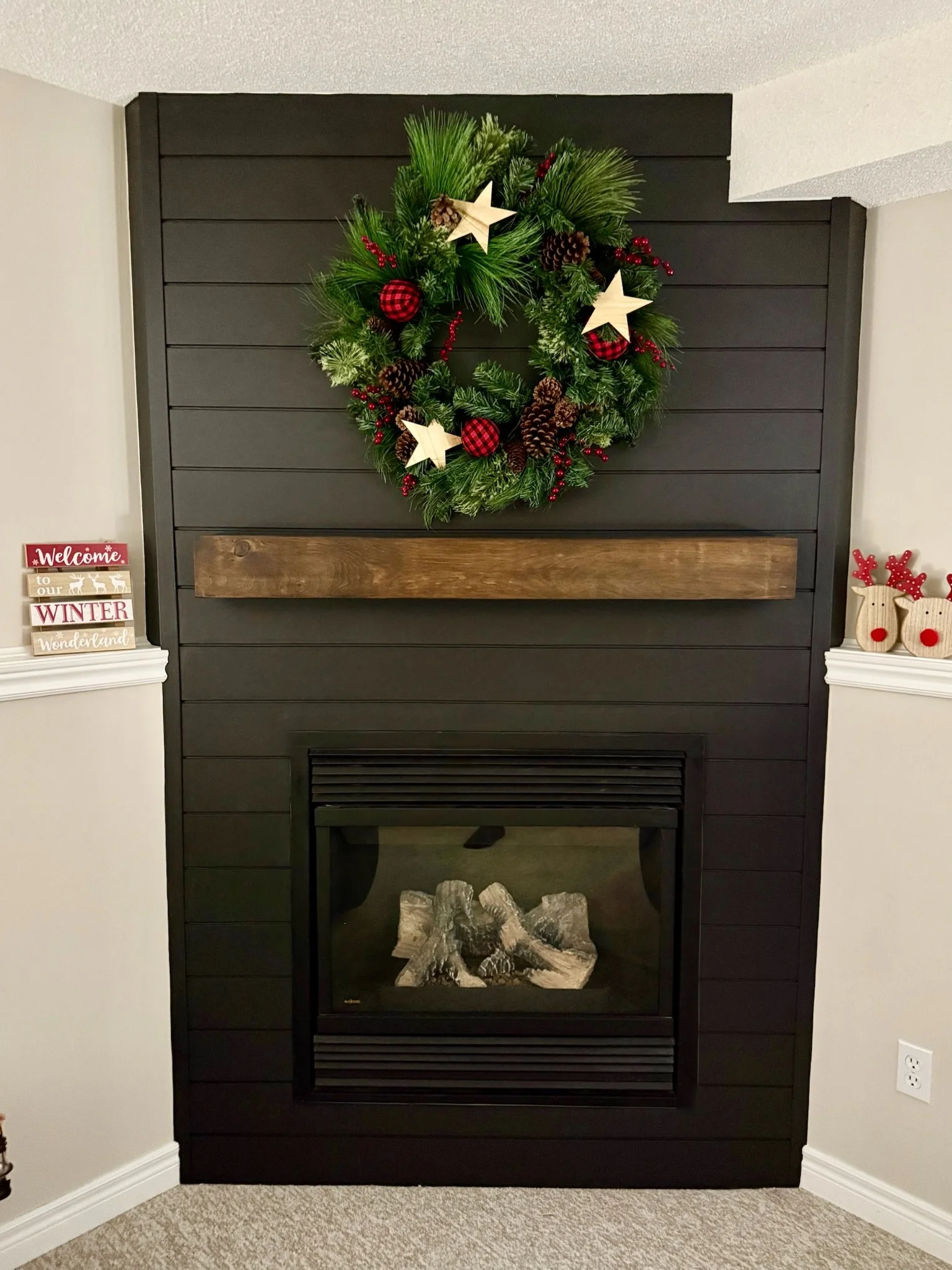 Black electric fireplace with a wooden mantel and a Christmas wreath hanging above it, decorated with pine cones, red berries, stars, and plaid-patterned ornaments.