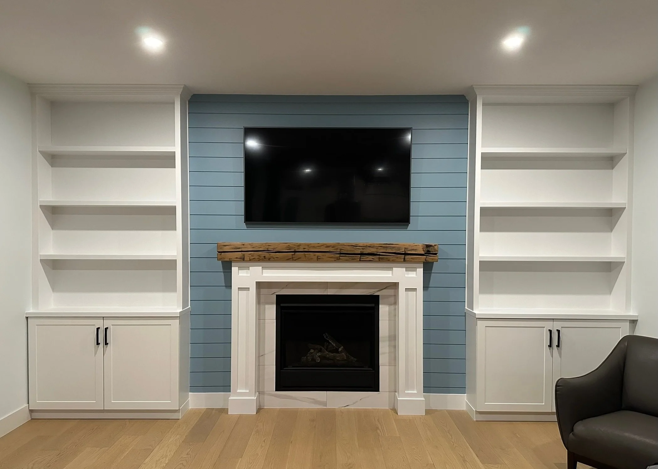 Living room with white bookshelves on either side of a fireplace with a wooden mantel. A flat-screen TV is mounted above the fireplace, which has a black interior. The wall behind the TV is painted blue with horizontal paneling.