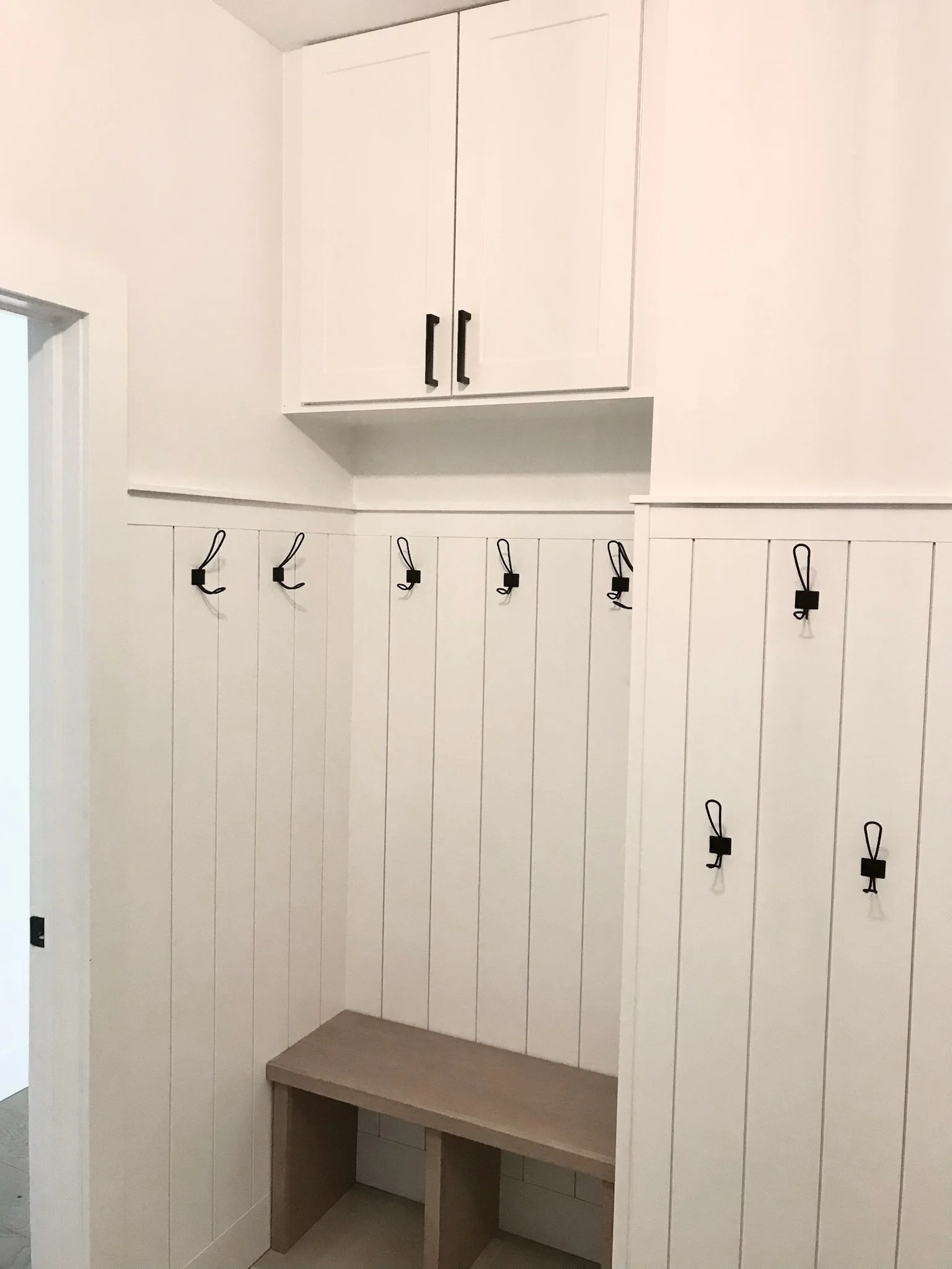 Corner of a mudroom with white shiplap walls, black hooks with straps, a small built-in bench with a wooden top, and a white cabinet with black handles above the hooks.