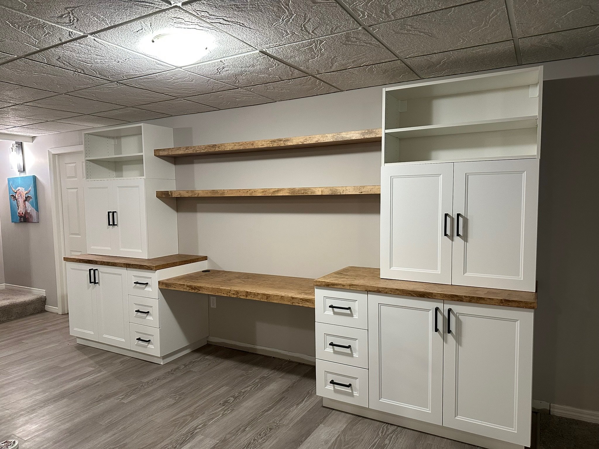 A room featuring white cabinets with black handles and natural wooden shelves. The ceiling is textured with a ceiling light. A small painting of a cow hangs on the wall.