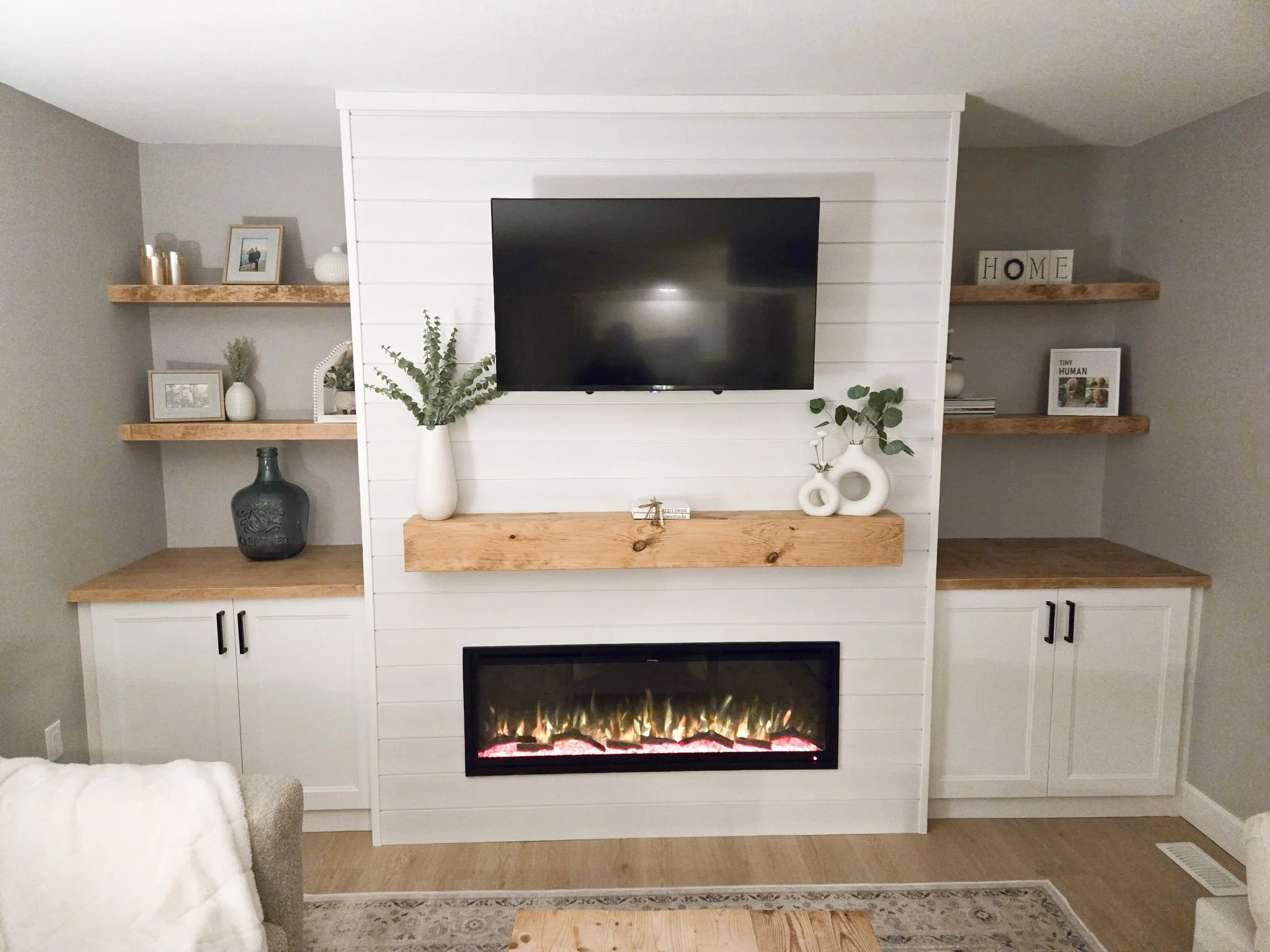 Living room wall with a white built-in entertainment center, a mounted flat-screen TV, a fireplace below, knotty wood mantel, and open wooden shelves on either side decorated with picture frames, vases, and plants.