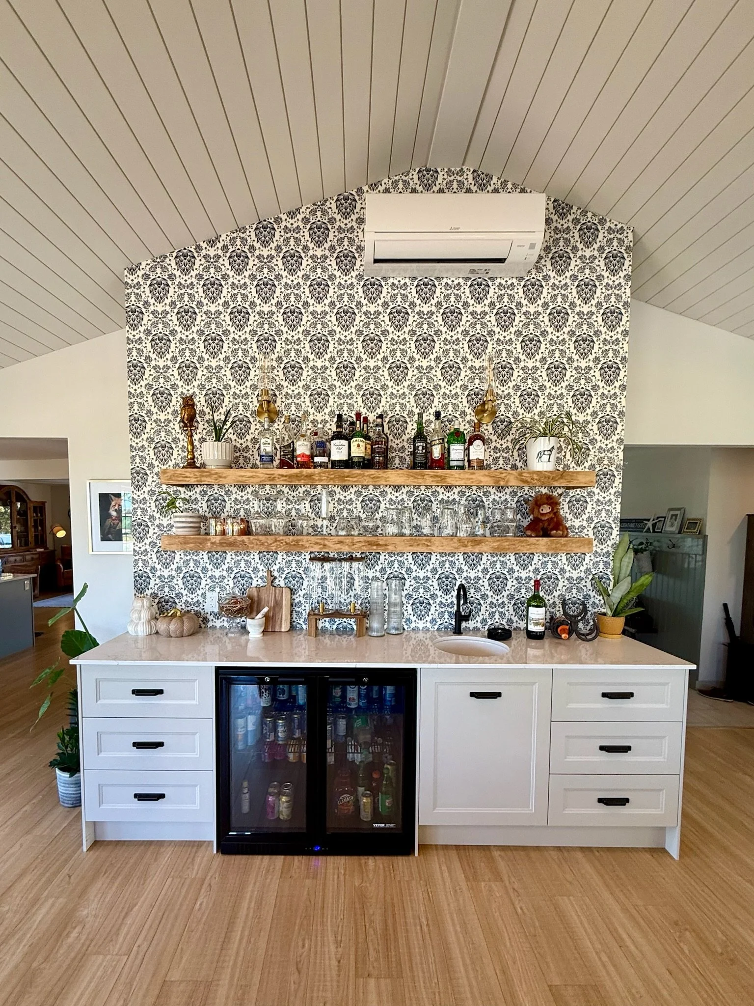 A modern kitchen bar area with white cabinetry, a quartz countertop, wooden shelves and a wine fridge, decorated with plants, and various bottles of alcohol. There is patterned wallpaper behind the bar.