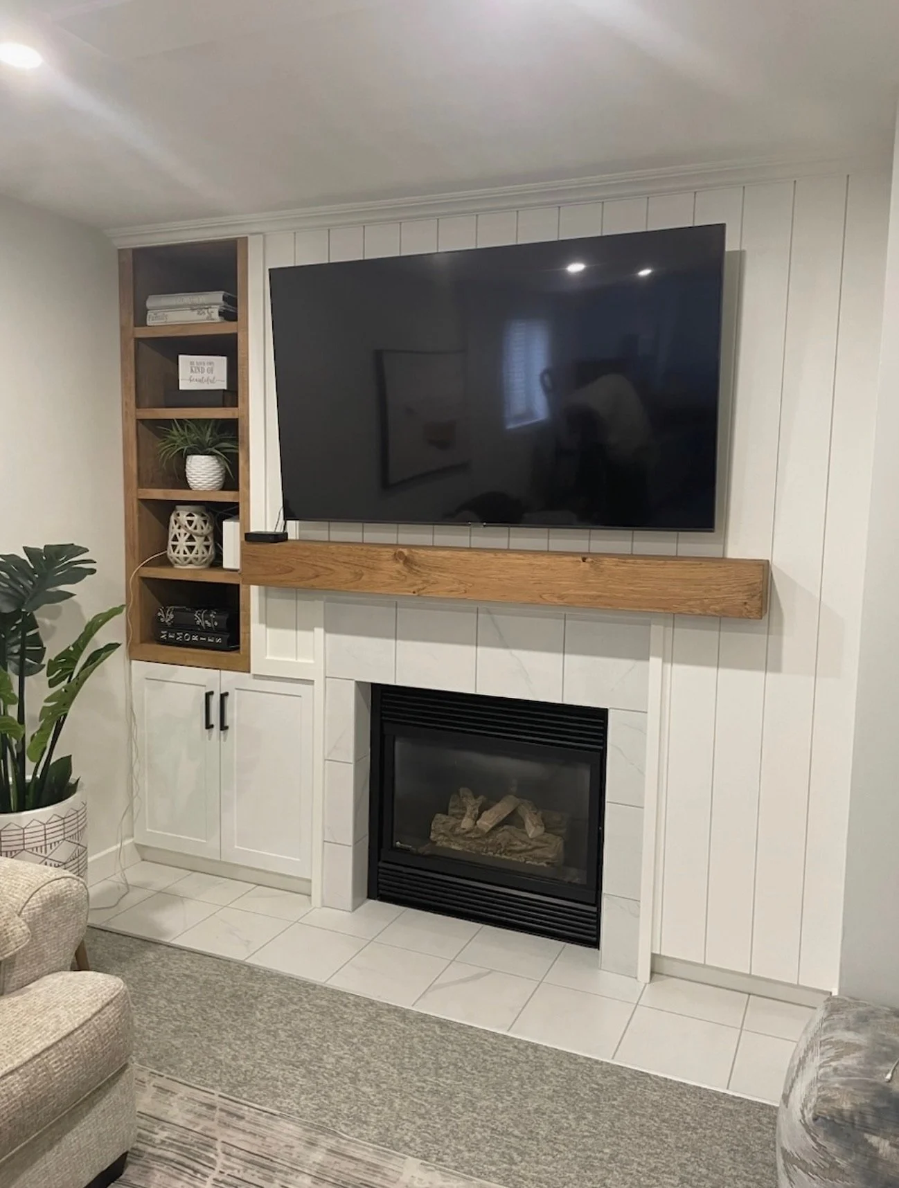 Living room with a wall-mounted flat-screen TV above a white fireplace, decorated with a wooden mantel and a white shiplap wall. To the left, a wooden bookshelf with books, a plant, and decorative items. A gray armchair and a patterned rug are partia