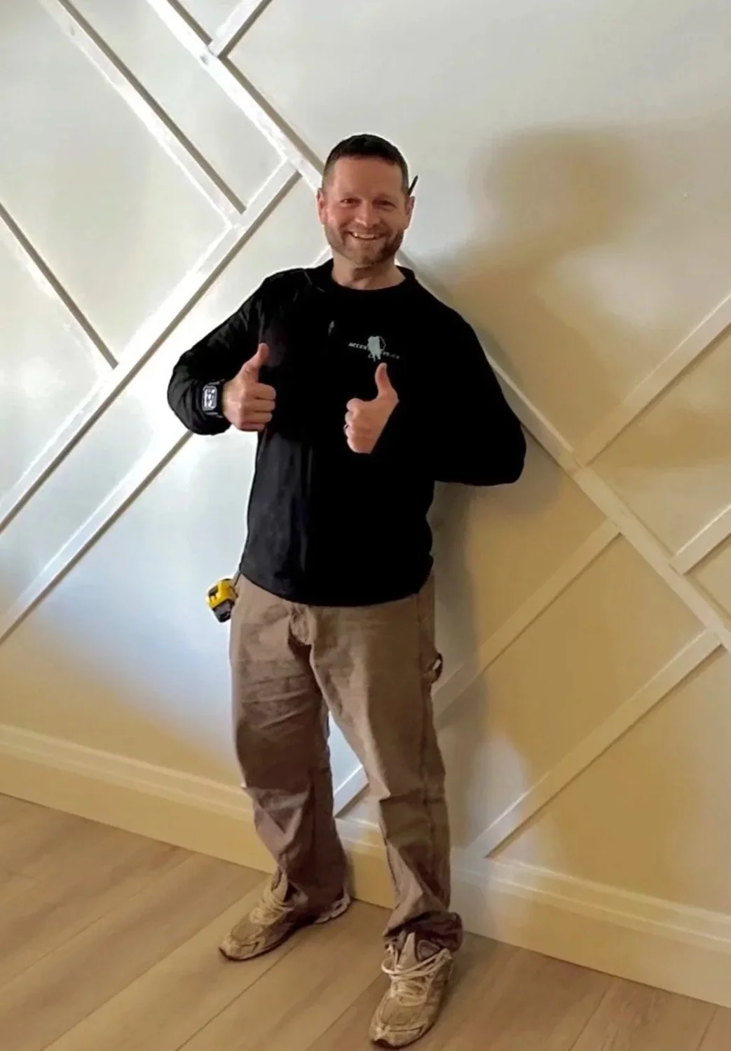 A man standing against a decorative wall, smiling, giving two thumbs up, wearing a black jacket, beige cargo pants, and beige sneakers.