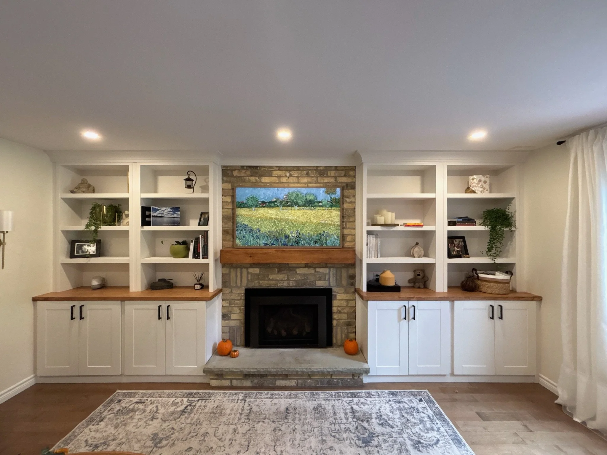 Living room with built-in white shelves around a brick fireplace, a landscape painting above the fireplace, pumpkins on the hearth, and a patterned rug on a wooden floor.