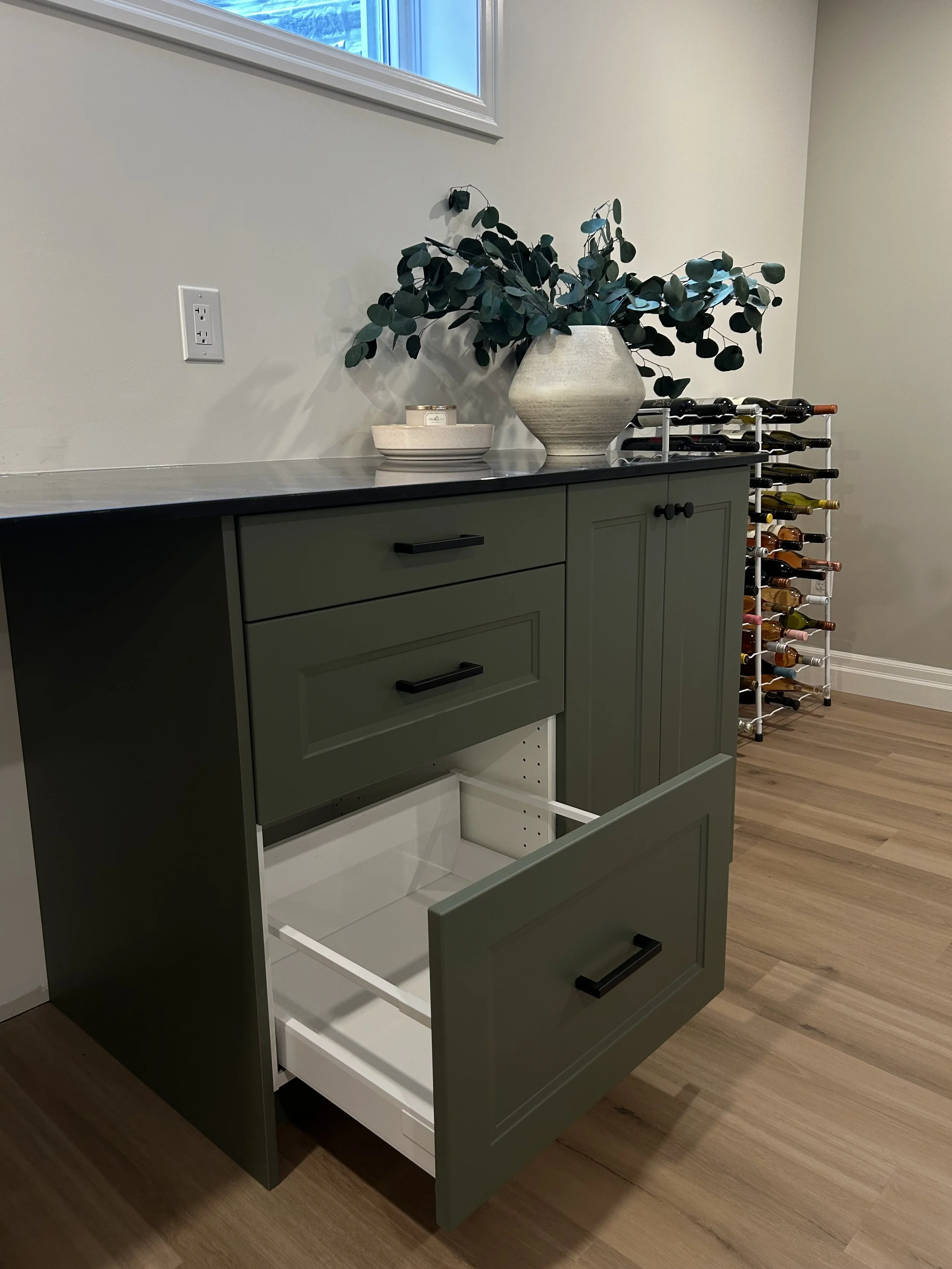 An unfinished green kitchen cabinet with open drawers, a white interior, and black handles, next to a wine rack filled with bottles, against a wall with a window.