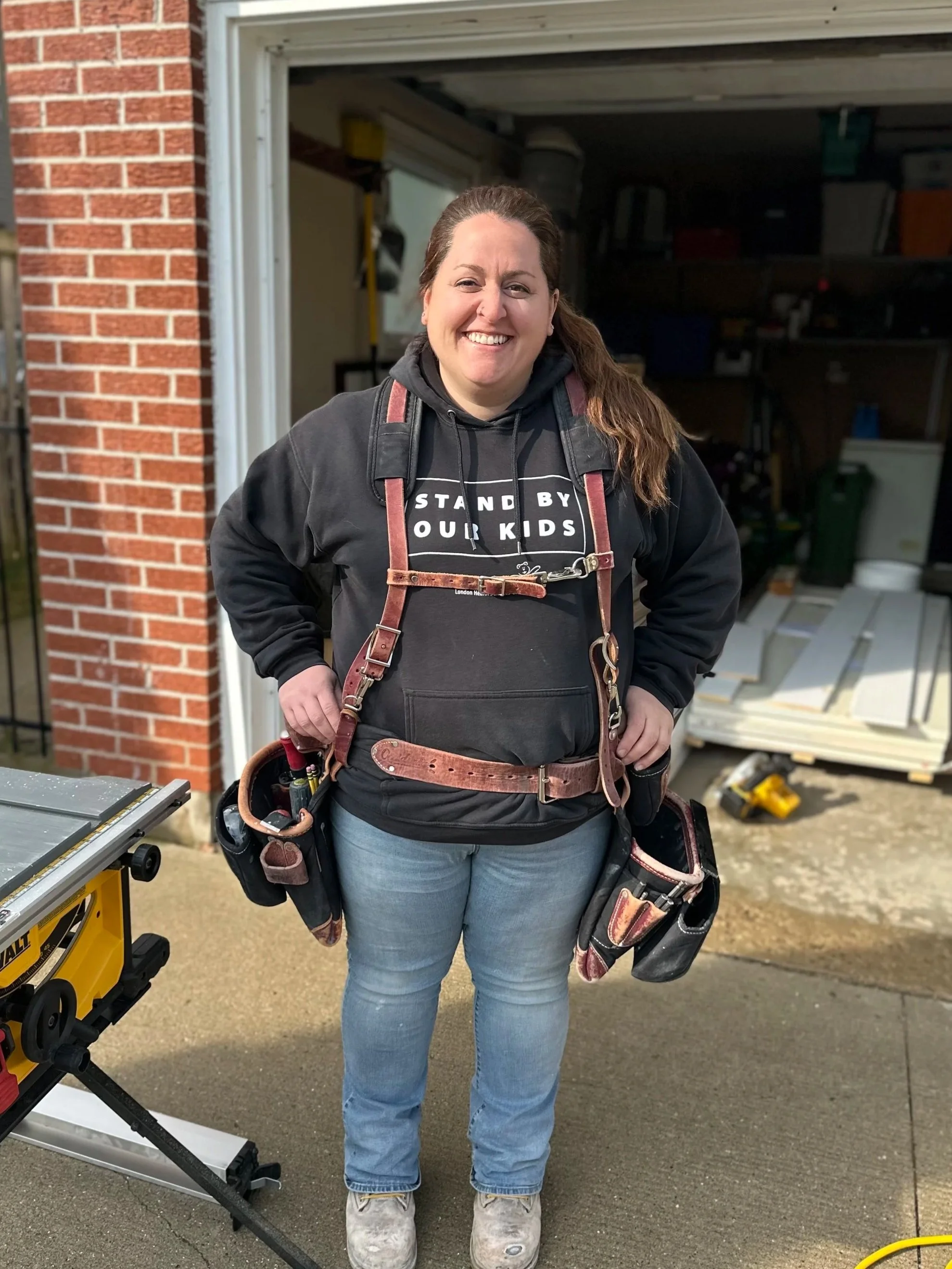 Smiling woman with a tool belt and backpack standing in front of an open garage and a workbench with tools.