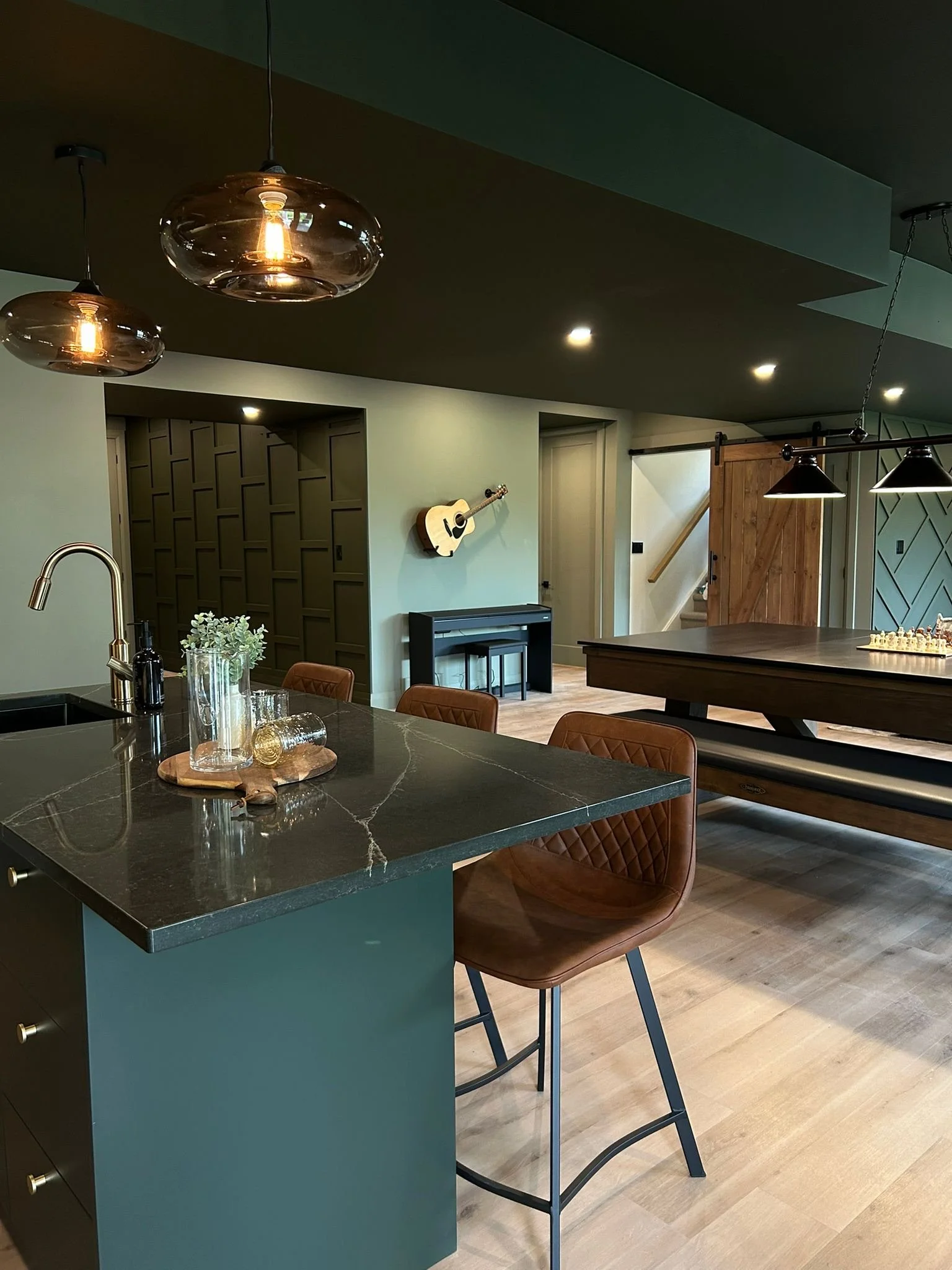Modern kitchen with black quartz island, brown quilted chairs, light wood flooring, hanging glass pendant lights, a guitar mounted on the light green wall, and a sliding barn door.