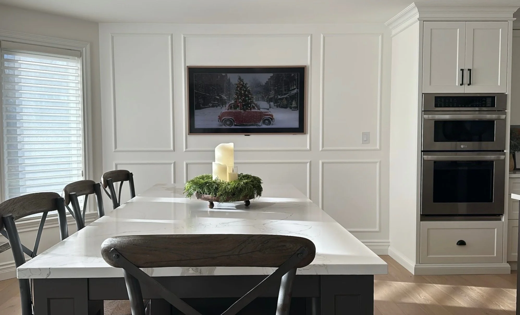 Modern kitchen with white cabinetry and stainless steel double oven, dining table with a white marble top, decorated with a green wreath and candles, windows with white shutters, and the picture frame moldings surround the TV.