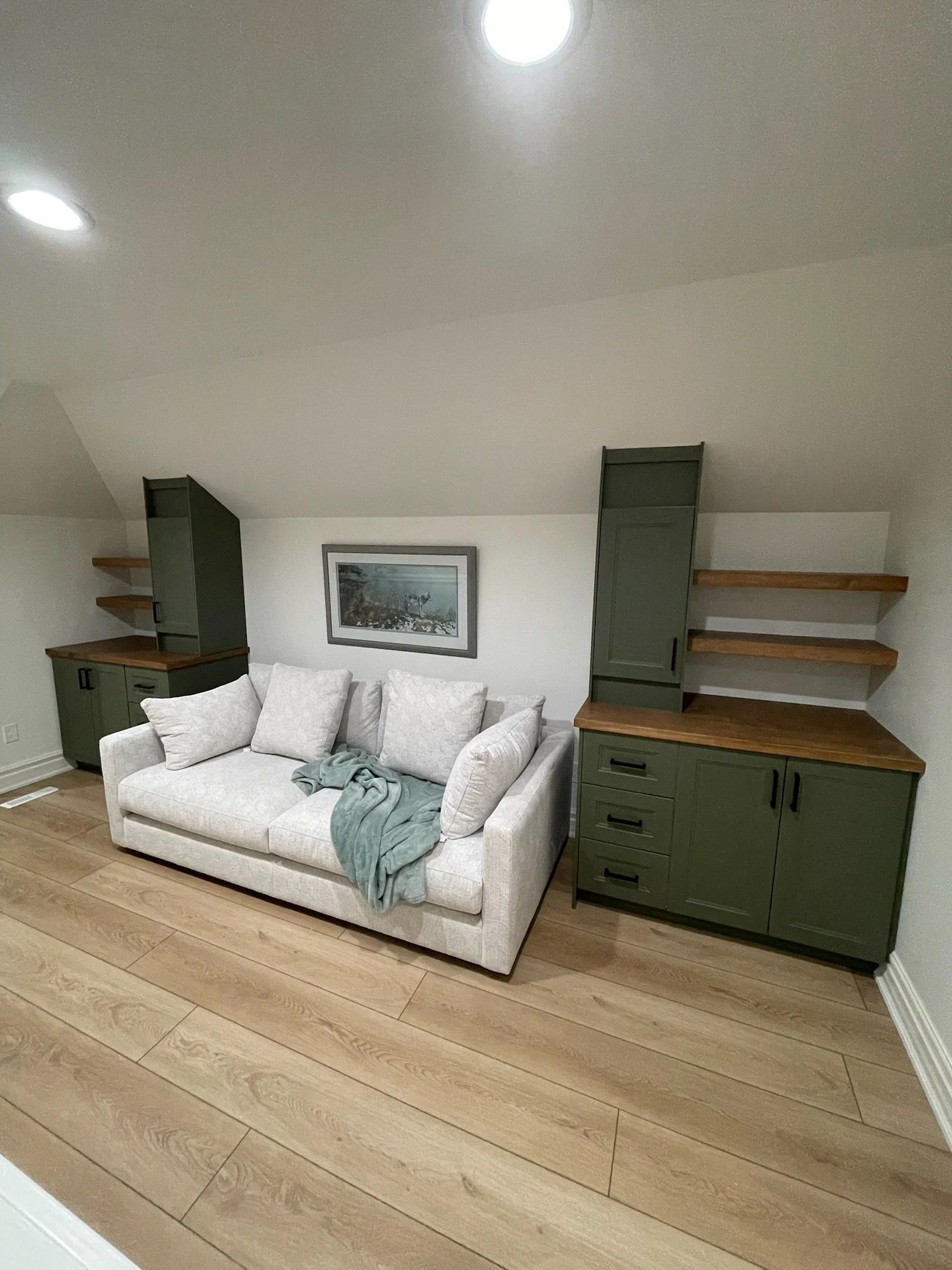 Living room with light-colored sofa, green built-in cabinets with wooden countertops, floating shelves, framed picture on wall, wooden plank flooring, and ceiling lights.